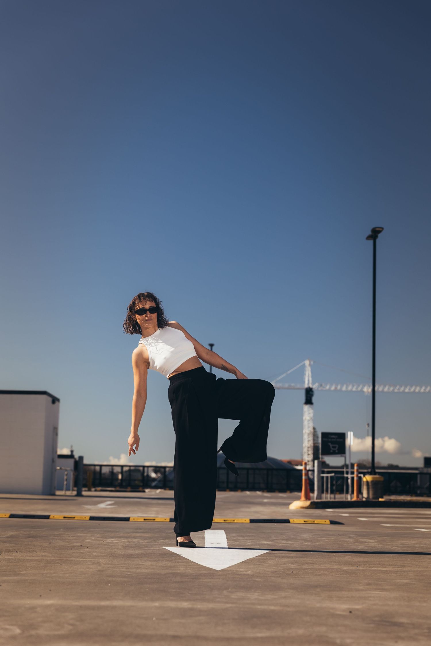 Woman in white crop top and black wide-leg pants posing dynamically on rooftop parking lot under blue sky.
