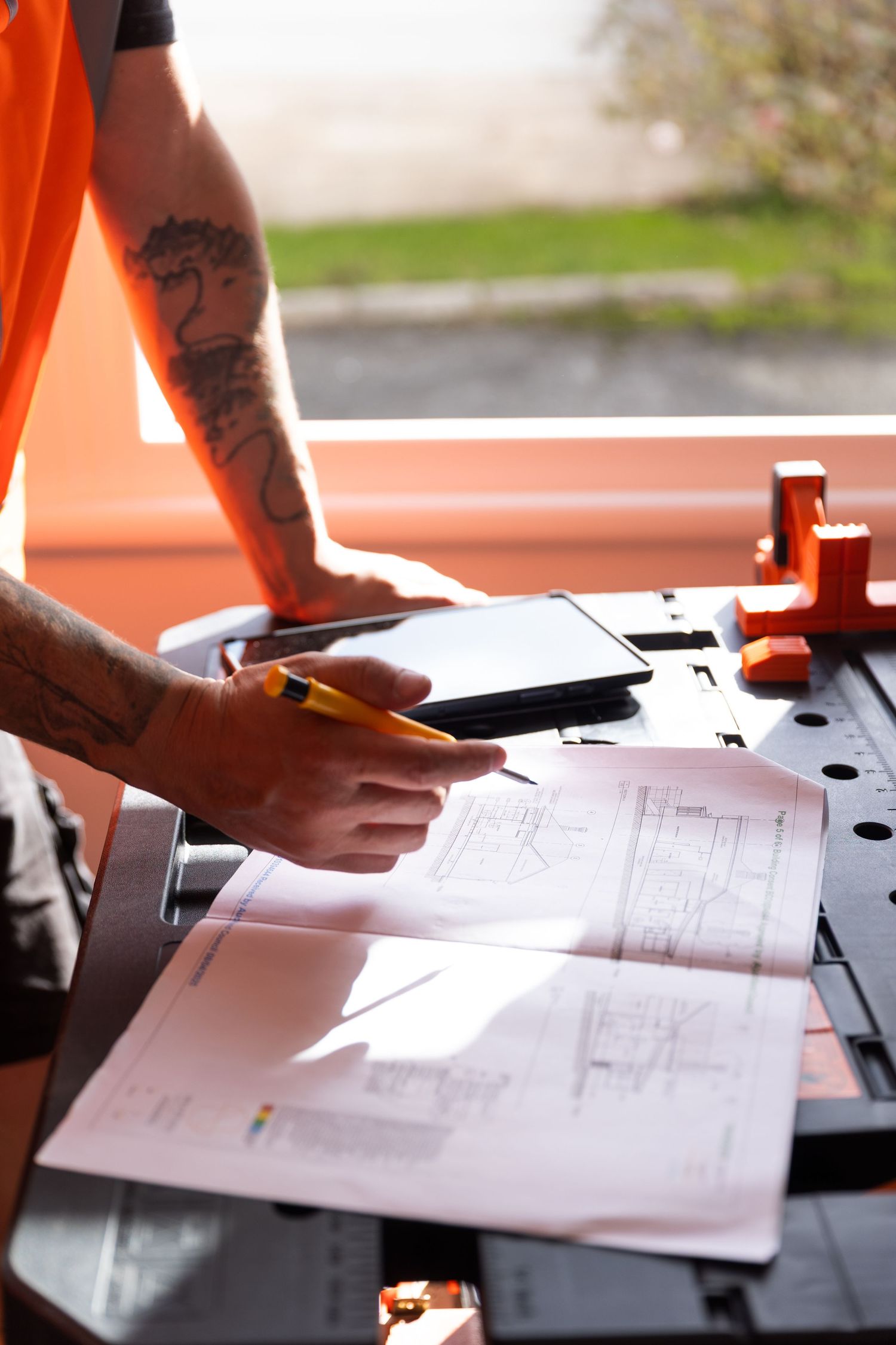 Tattooed construction worker holding pencil reviewing architectural blueprints on workbench.