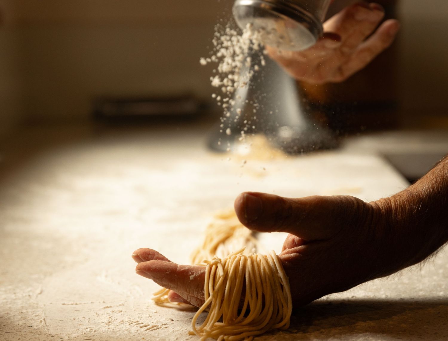 Hand holding a nest of fresh spaghetti while flour is dusted over it from above.