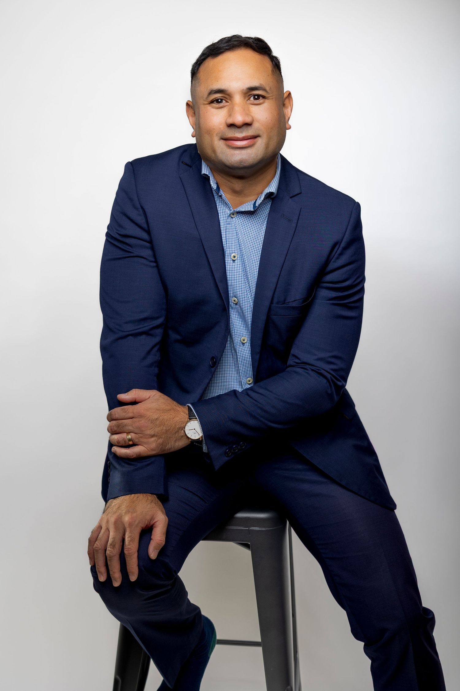 Professional man in navy blue suit sitting relaxed on a stool against a white background.