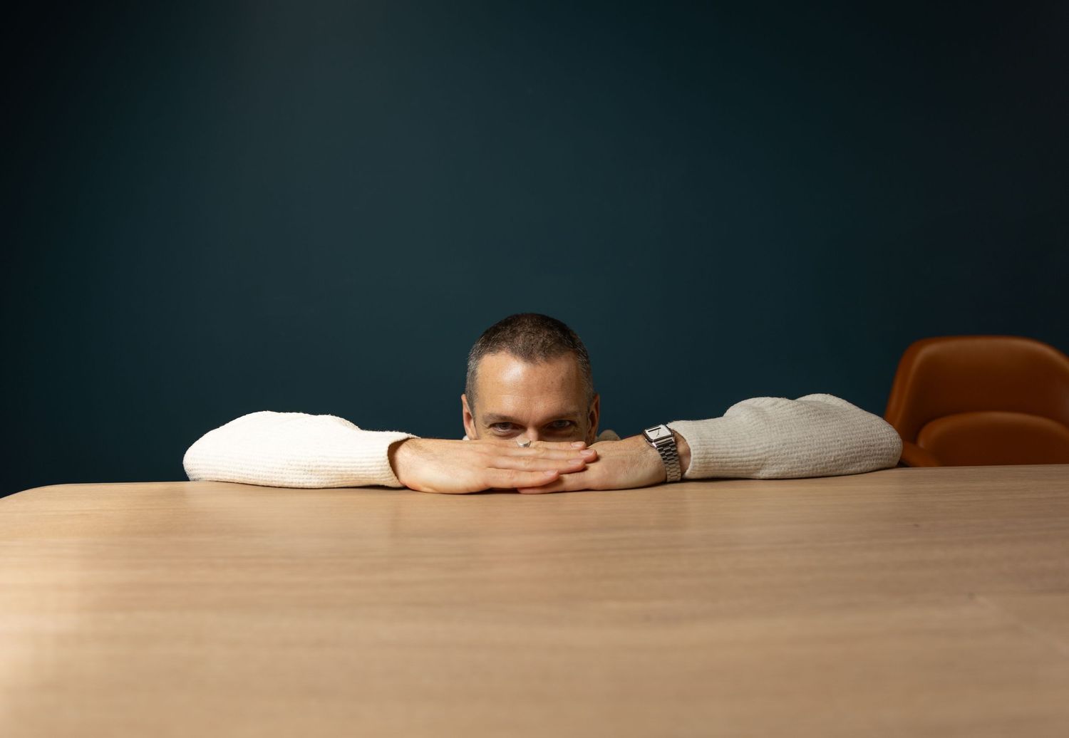 Man in a white shirt resting his chin on arms folded over a wooden table against a blue wall.