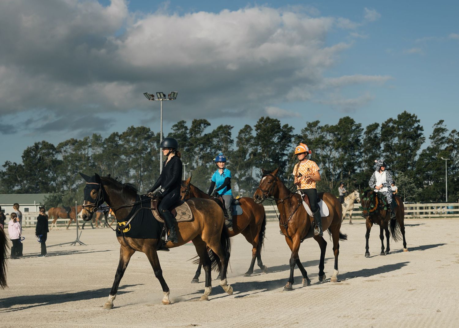 Four riders on horseback parade across a sandy arena under dramatic cloudy skies at an equestrian event.