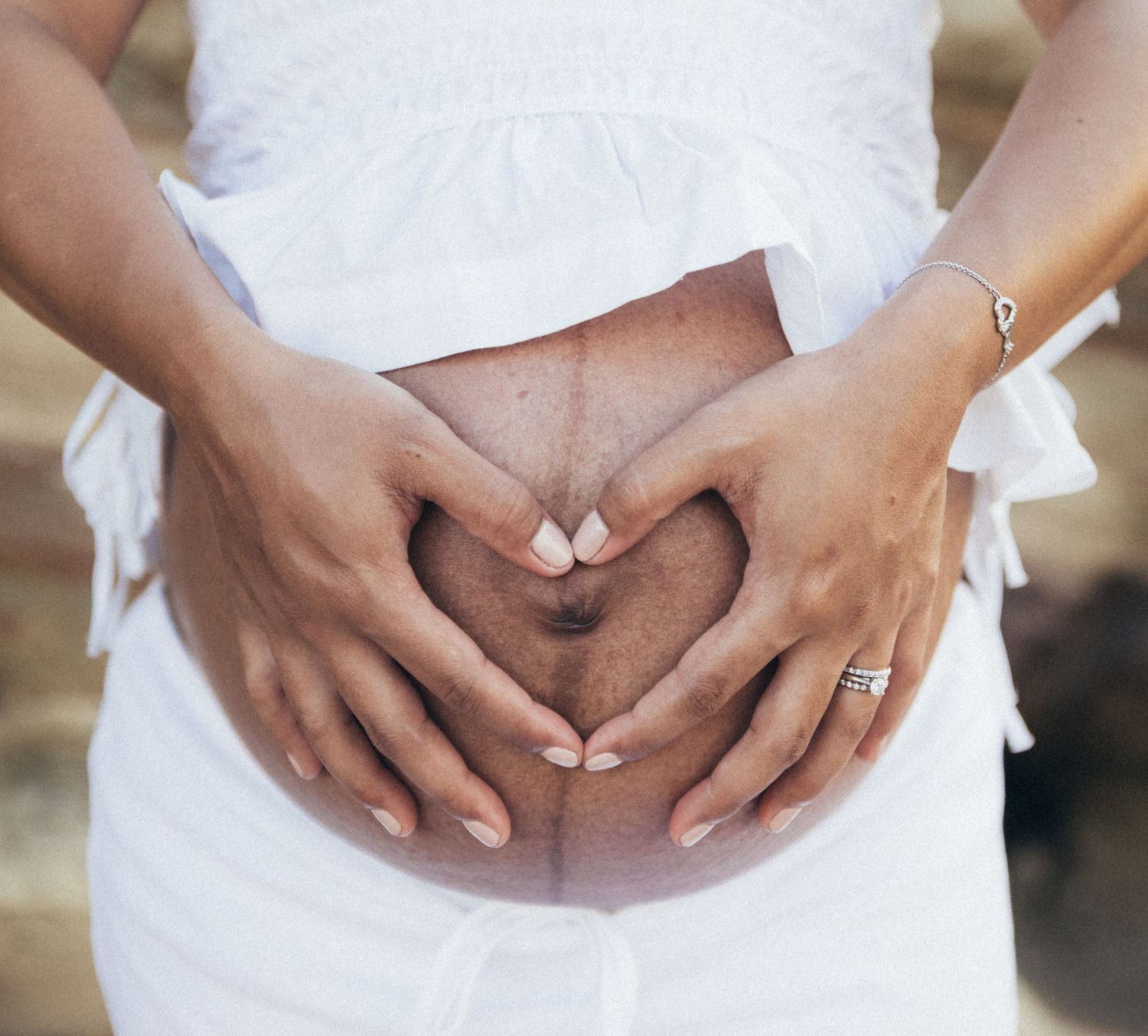 Pregnant woman in white dress forming heart shape with hands on baby bump, close-up maternity photo.