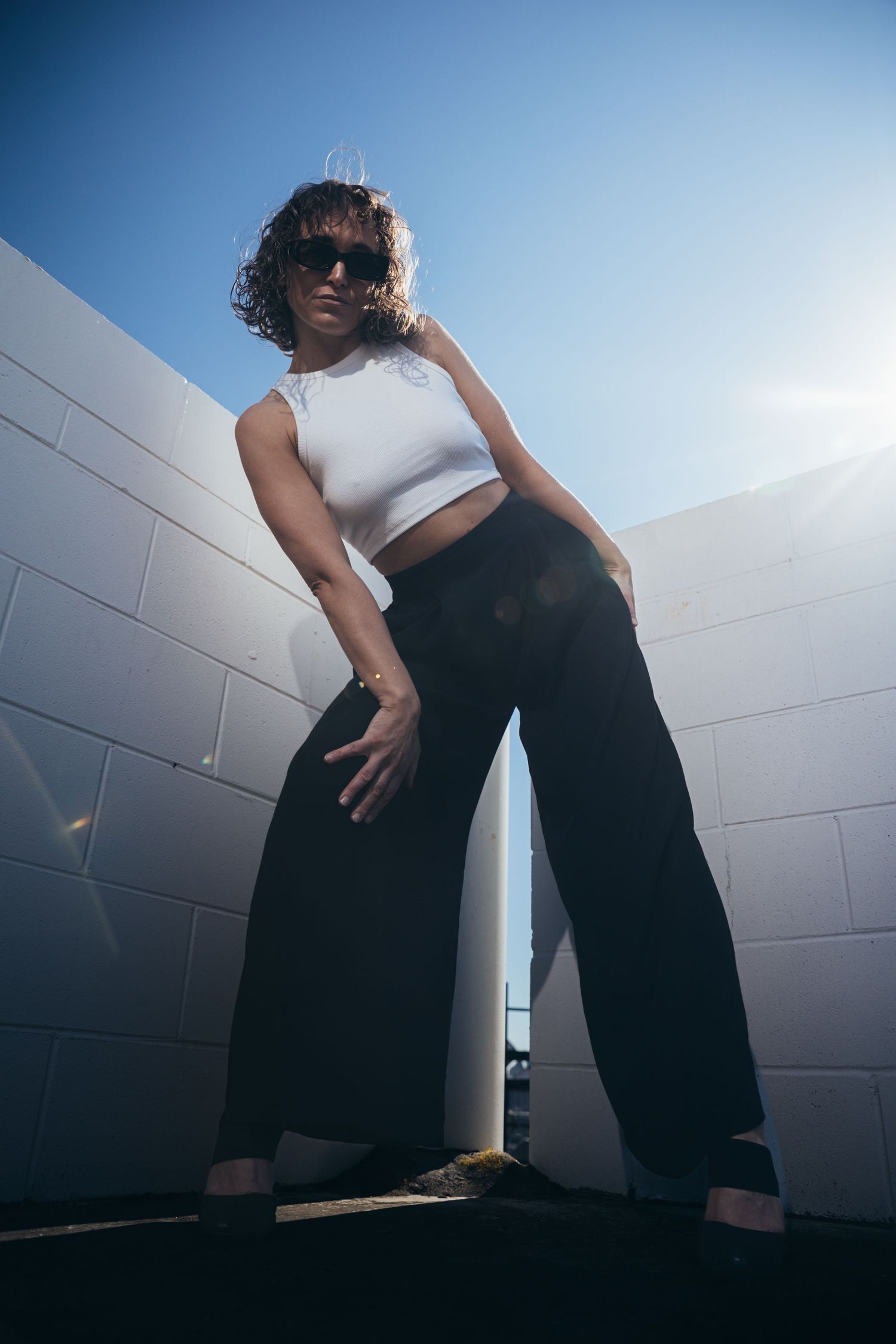 Woman in white crop top, black wide-leg pants and sunglasses posing against white brick wall under blue sky.
