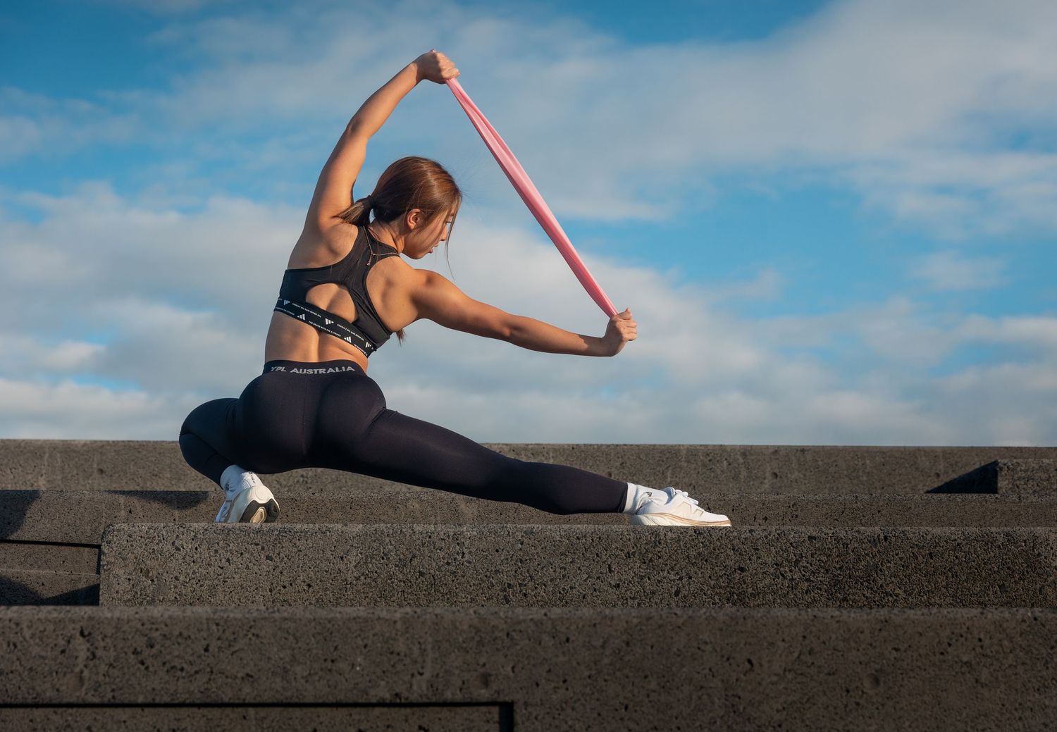 Athletic woman in black sportswear doing a deep lunge stretch with resistance band outdoors.