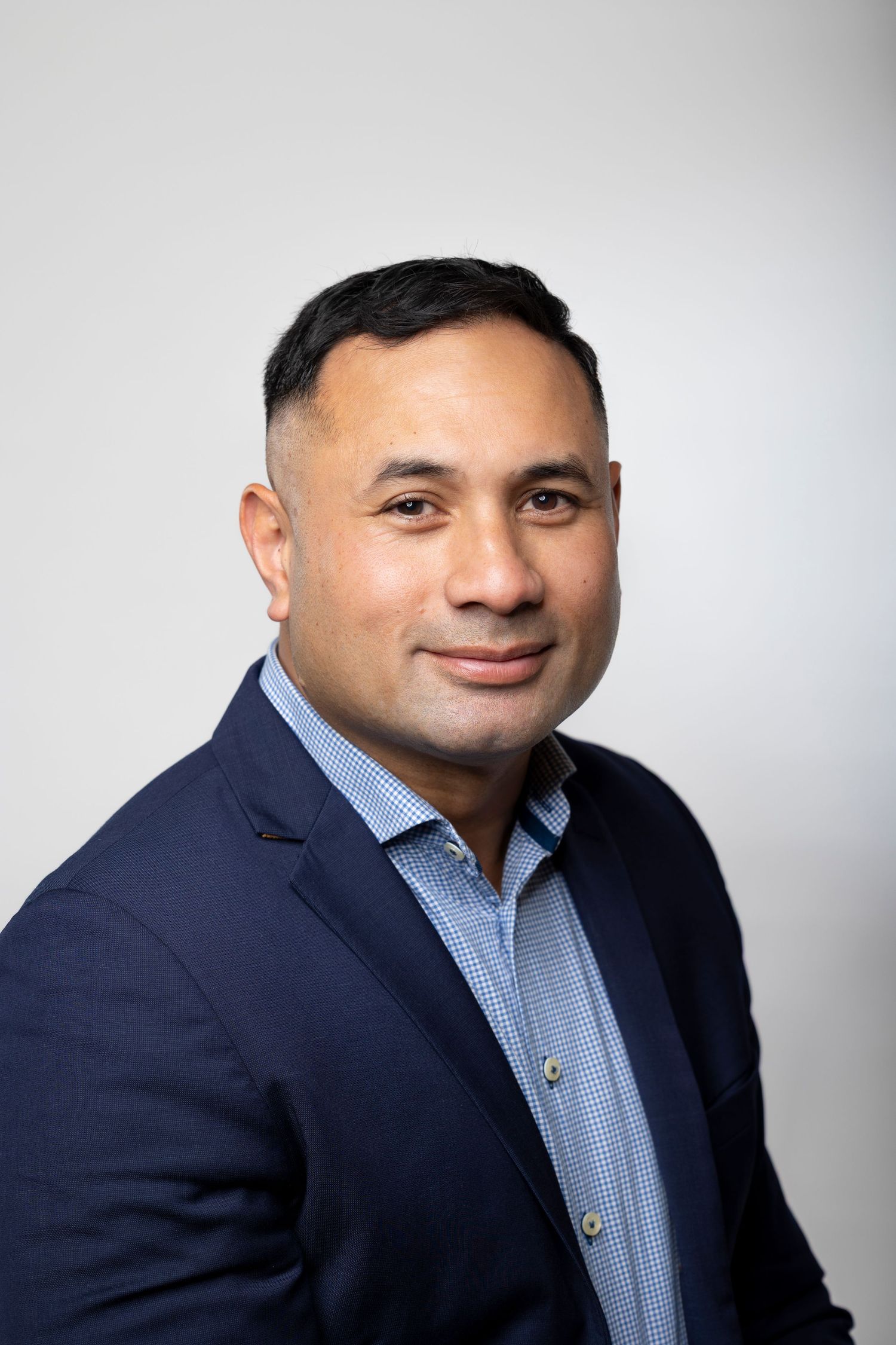 Professional man in navy suit and checkered shirt smiling in corporate headshot on gray background.