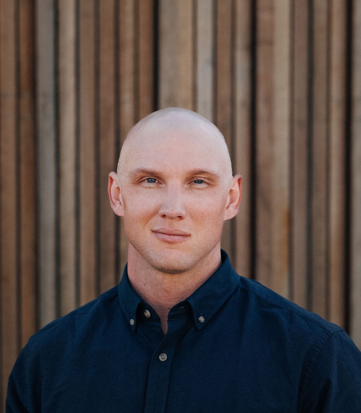 Bald man in navy blue button-up shirt smiling outdoors against wooden slat wall background.