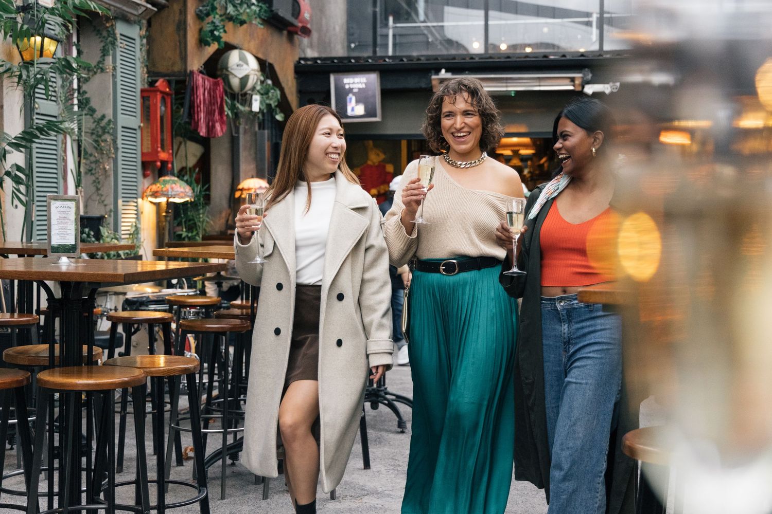 Three diverse women laughing and holding champagne glasses outside a restaurant patio.