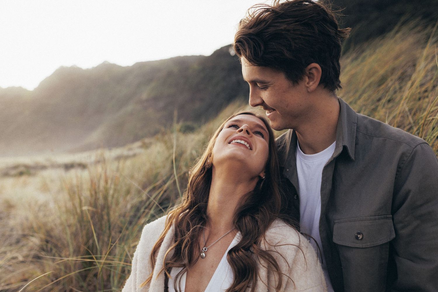 Smiling couple sharing a tender moment in coastal dunes with mountains behind them.