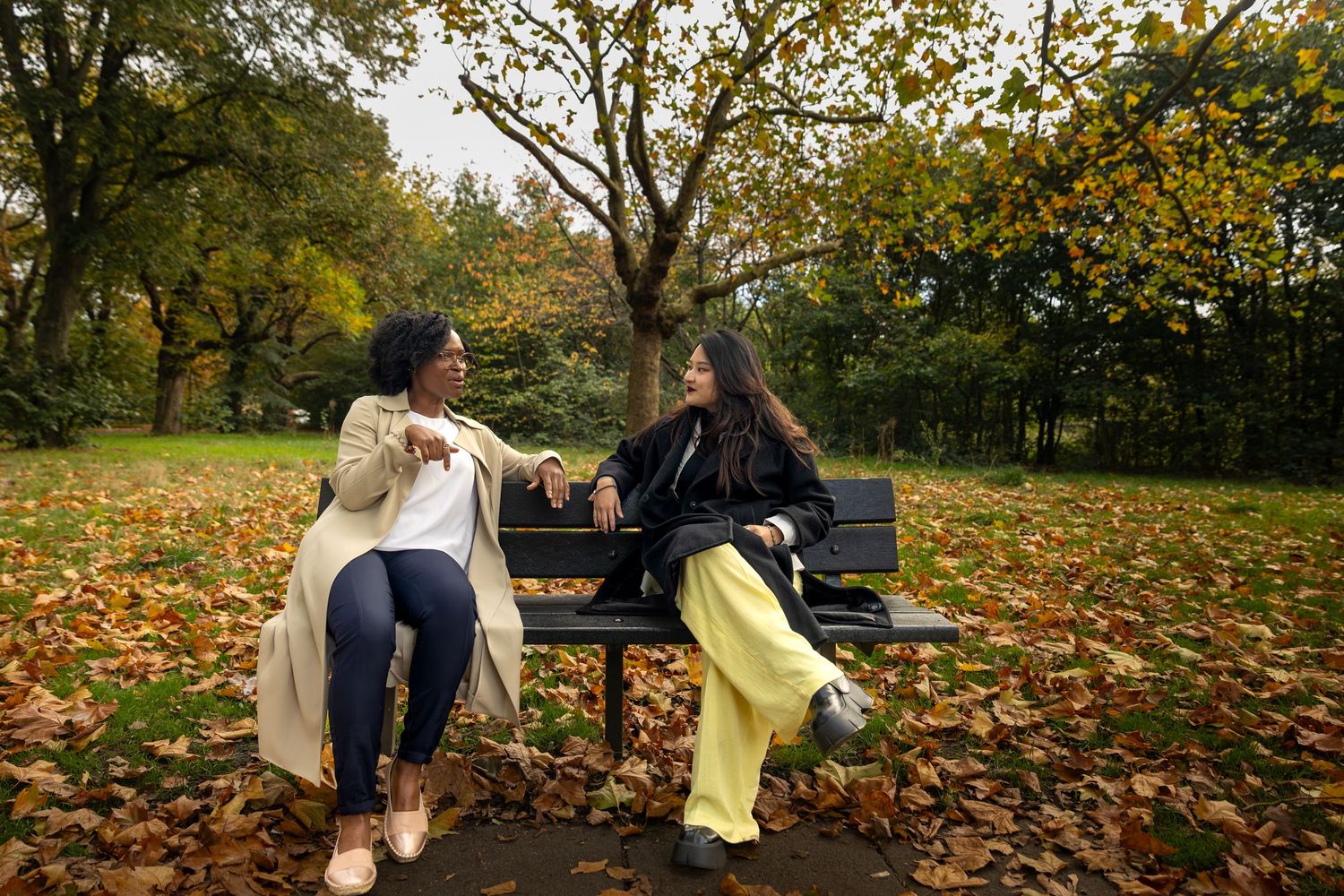 Two women sitting on a bench in an autumn park covered in fallen leaves, chatting together.