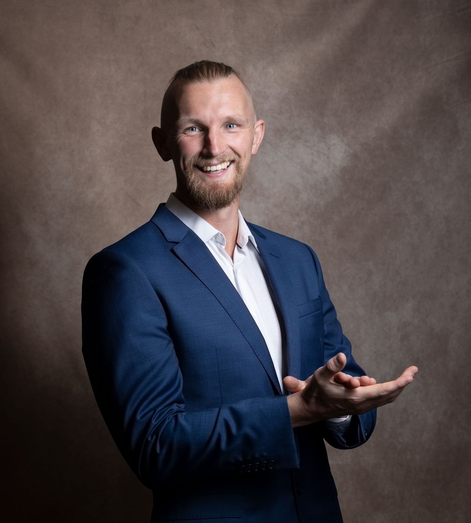 Smiling man in blue blazer gesturing with open hands against brown studio backdrop.