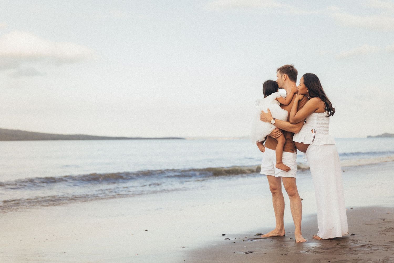 Family in white outfits embracing on a serene beach at dusk with calm ocean waves in the background.
