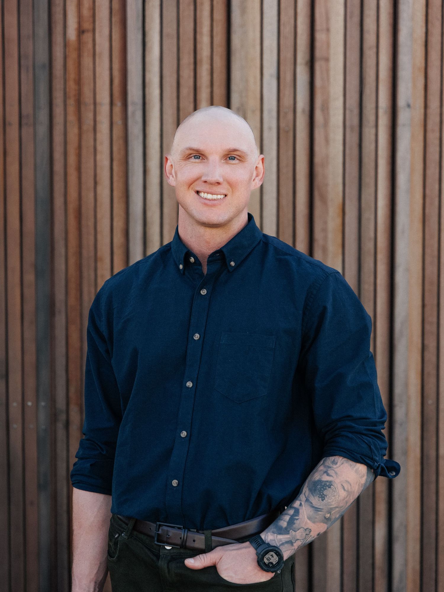 Smiling bald man with tattoos wearing a navy blue button-up shirt standing against a wooden slat wall.
