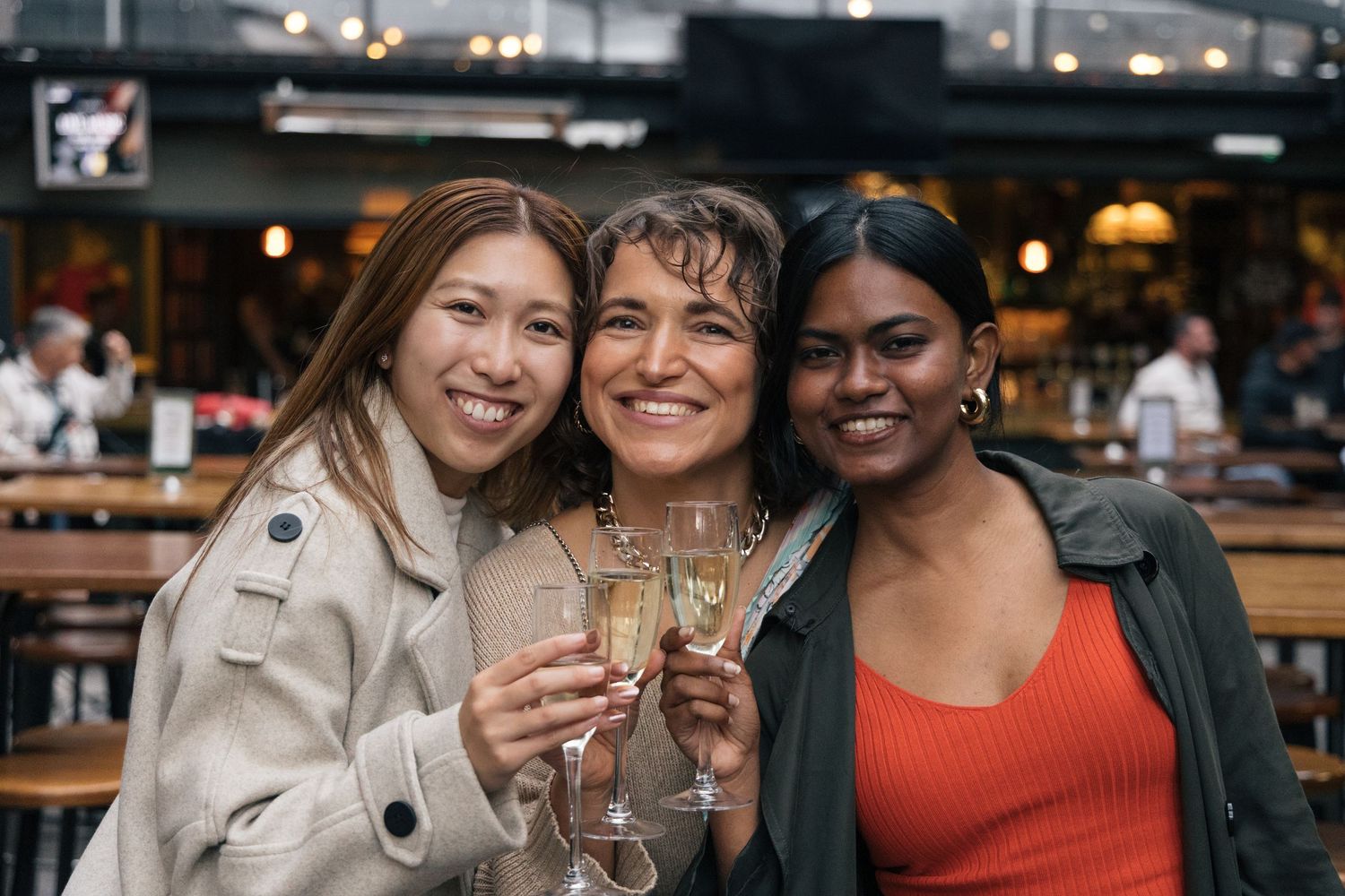 Three smiling diverse women posing together and toasting with champagne at an outdoor bar.
