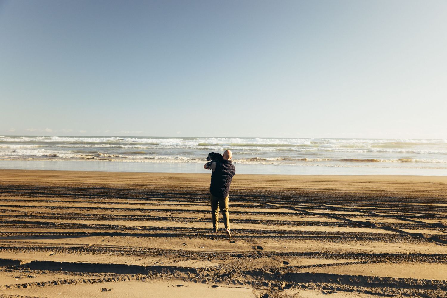 Person carrying dog while walking on wide sandy beach toward ocean waves in bright sunlight.