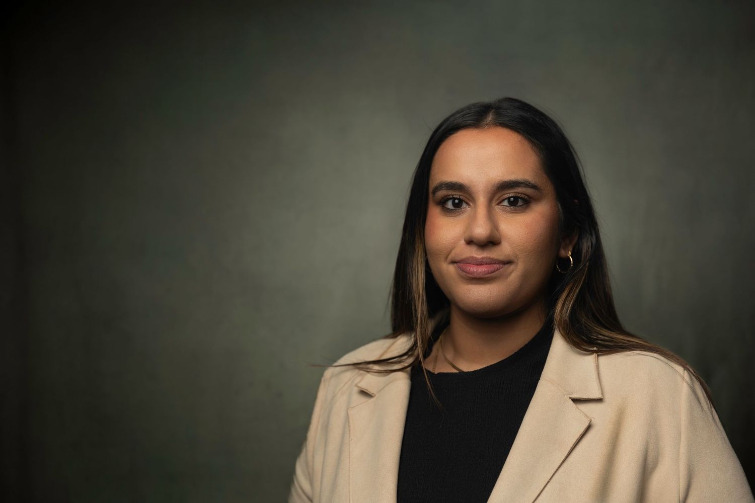 Young woman in beige blazer smiles confidently against dark green studio background.