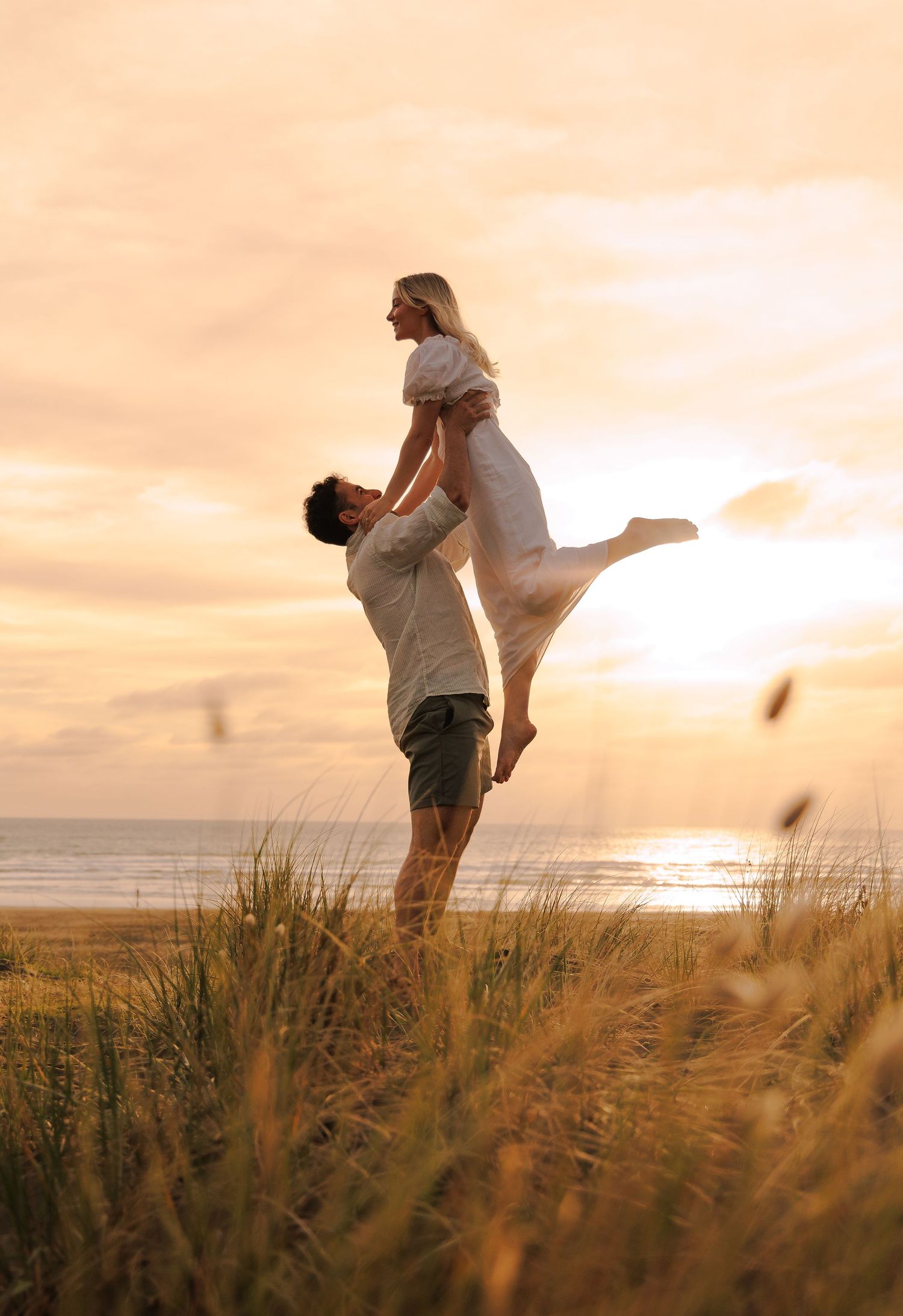 Man lifting woman in white dress at golden sunset beach, romantic couple photo in coastal grass dunes.