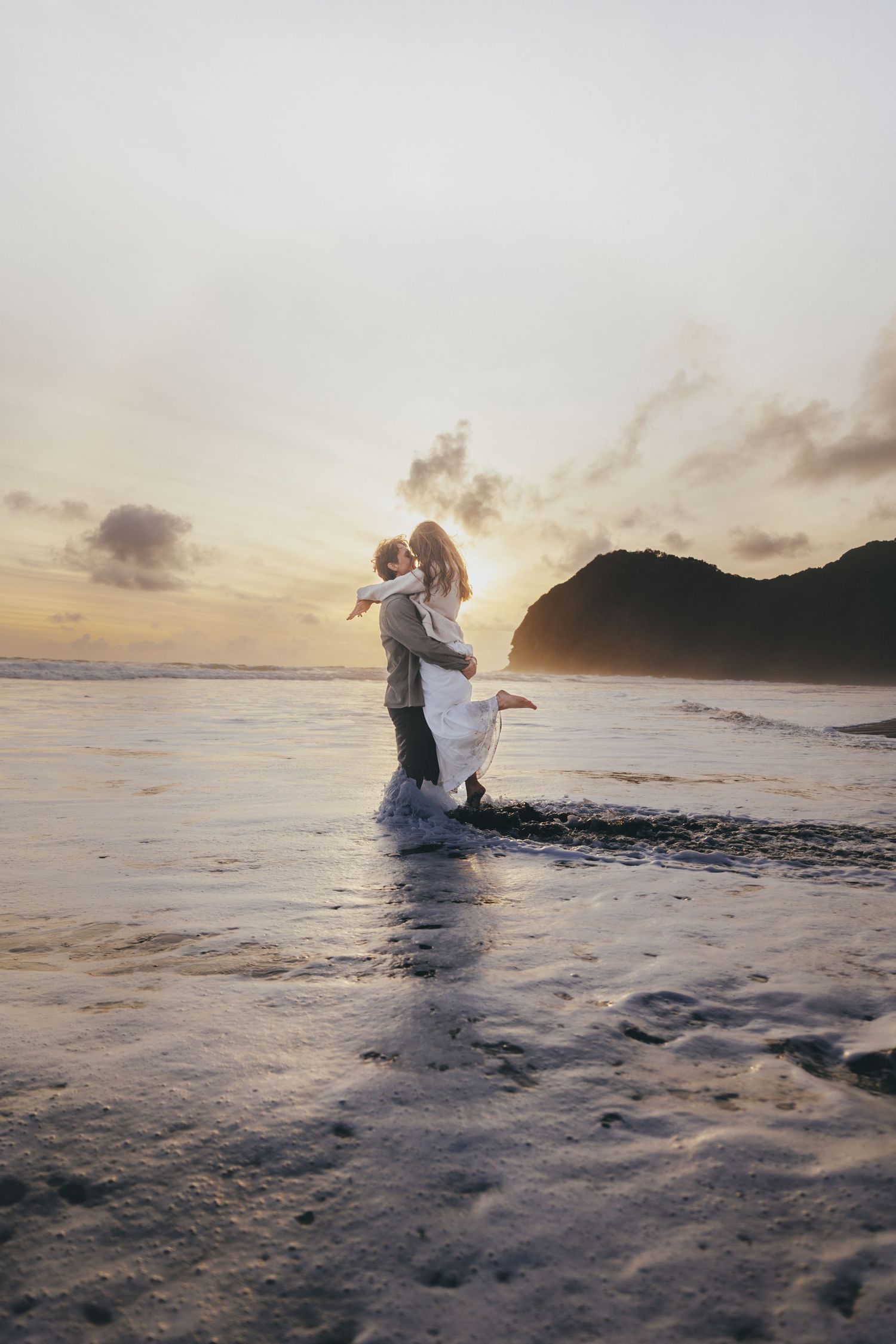 Couple embracing on a beach at sunset, waves washing ashore with a dramatic golden sky and coastal mountain backdrop.