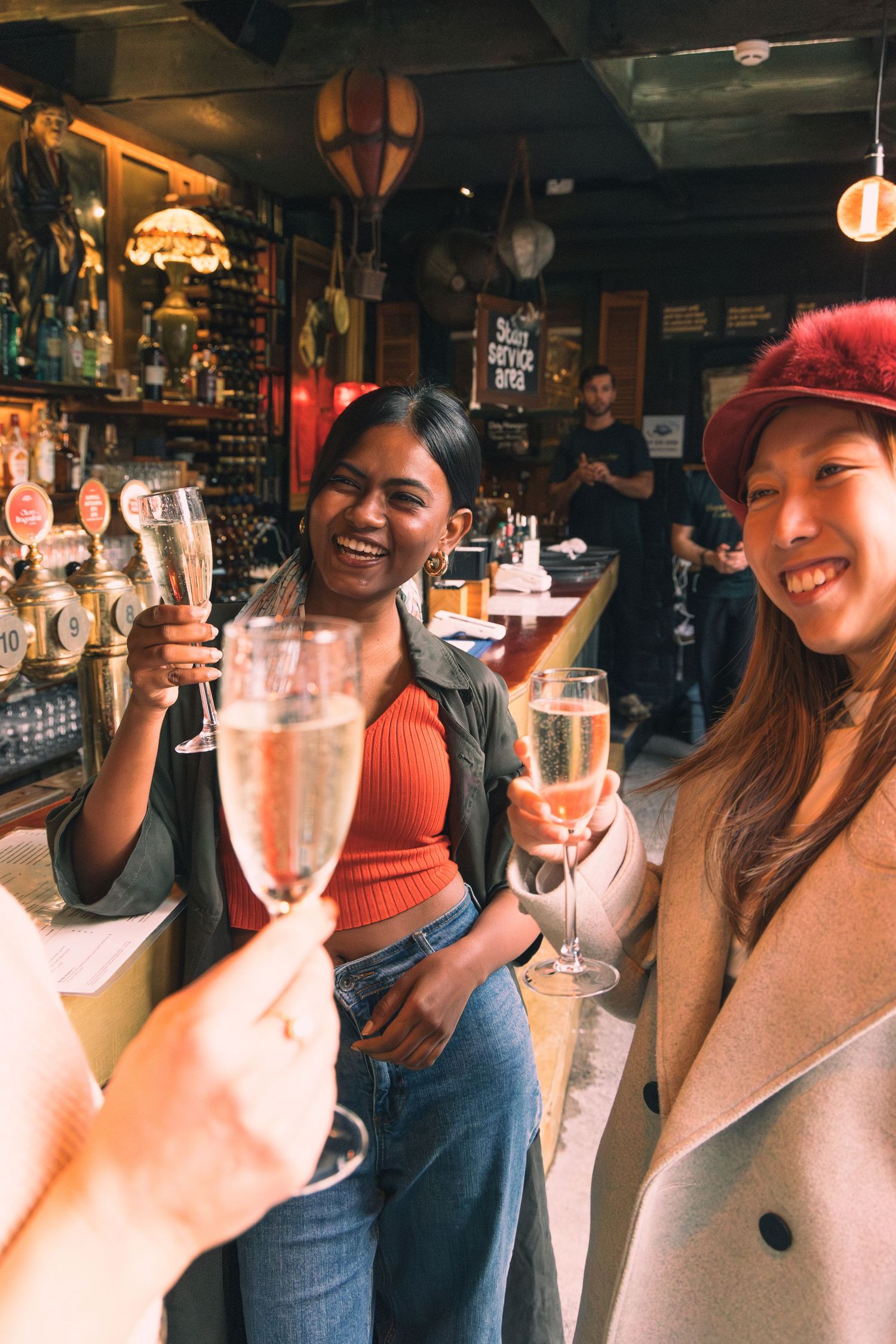 Two women smiling and toasting with champagne glasses at a warm, cozy bar.