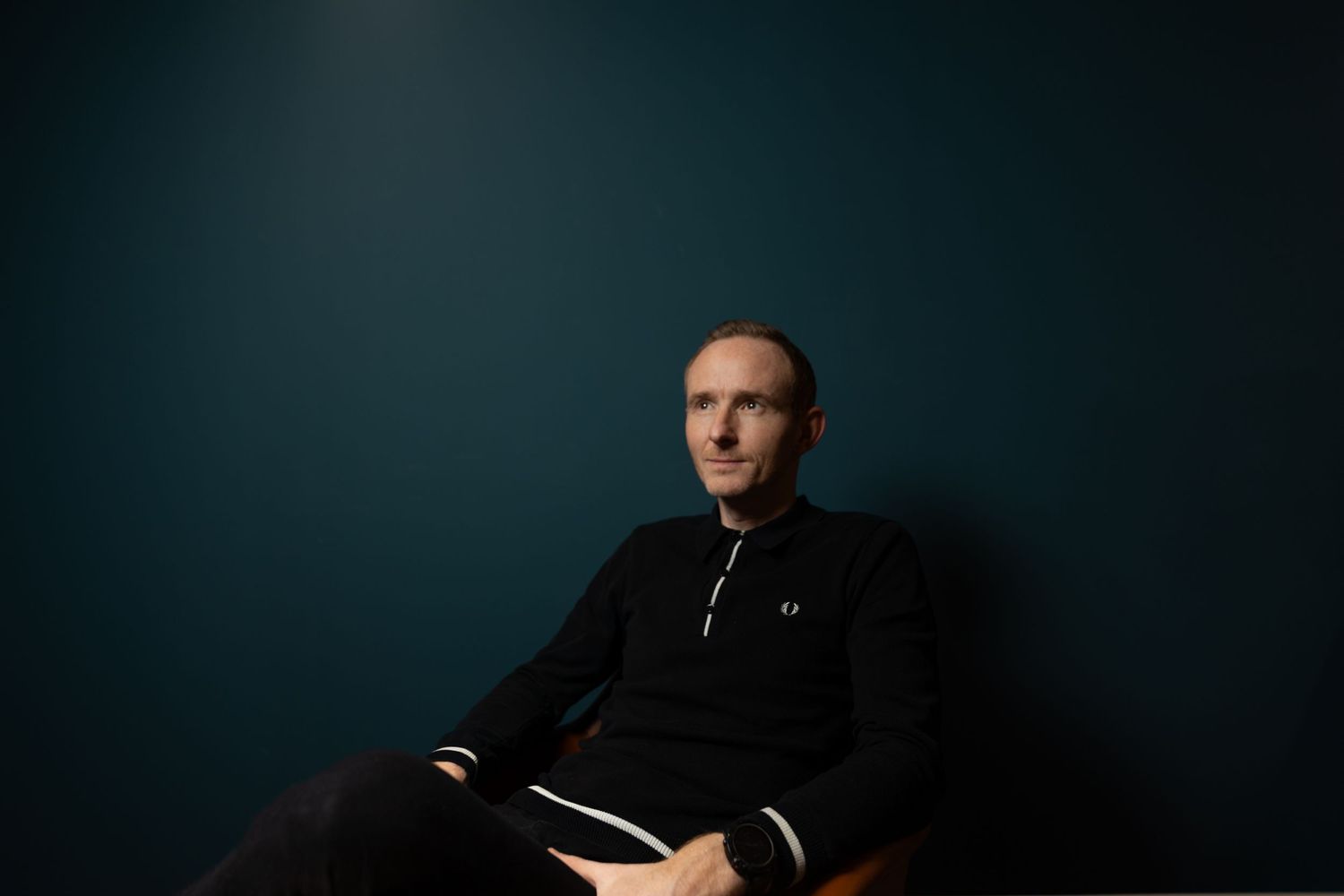 Calm man in a black Fred Perry tracksuit seated in a chair against a dark teal background.
