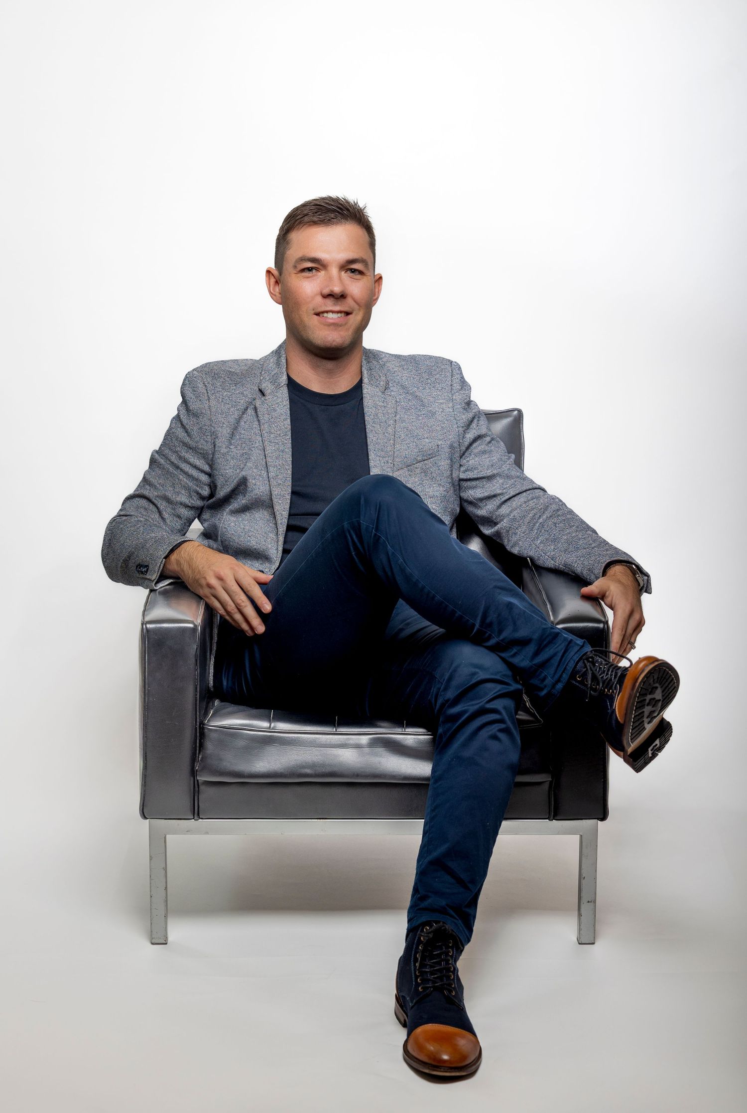 Relaxed man in grey blazer and jeans sitting in a leather chair against white background.