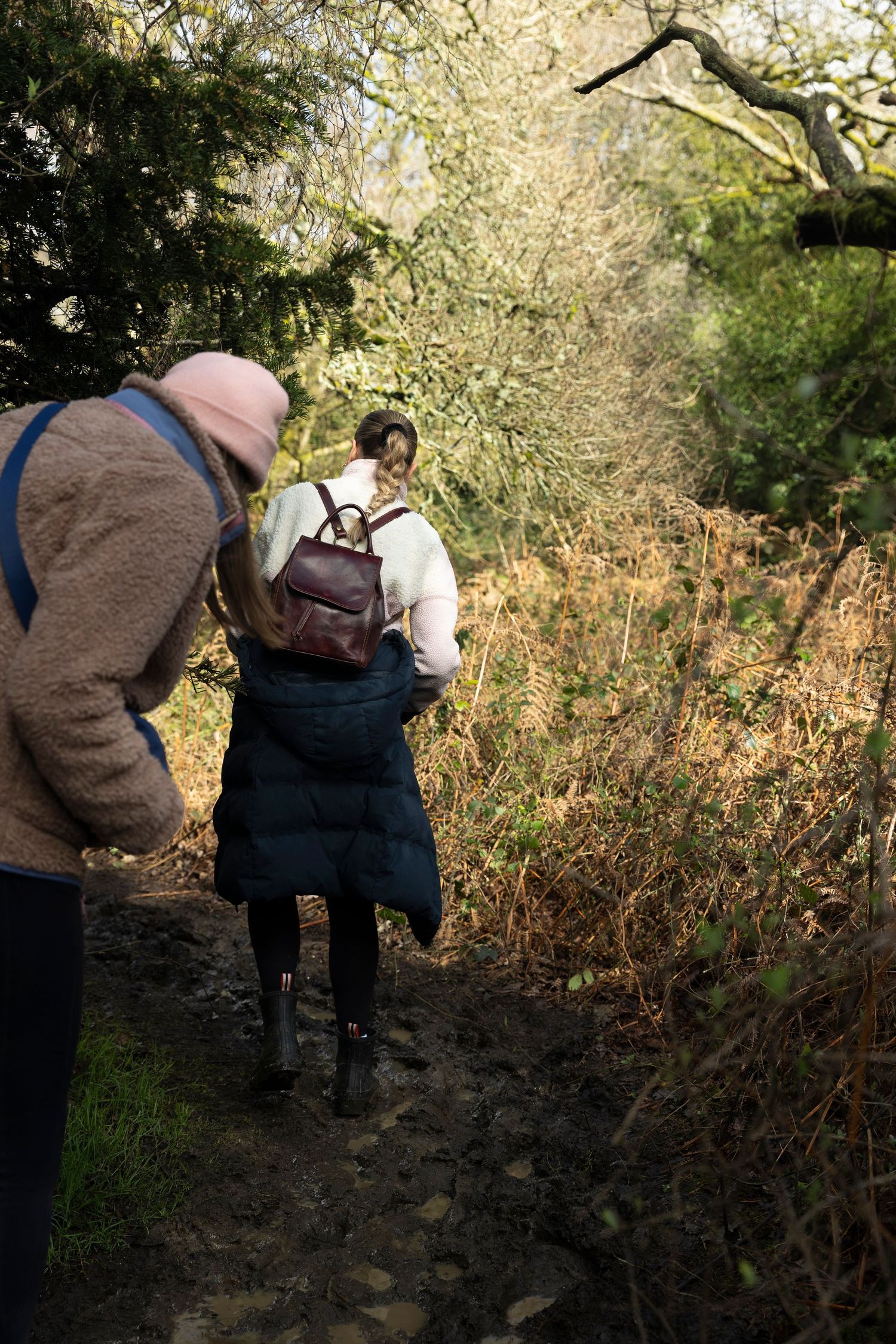 Two hikers walking along a narrow sunlit path through dense woodland.
