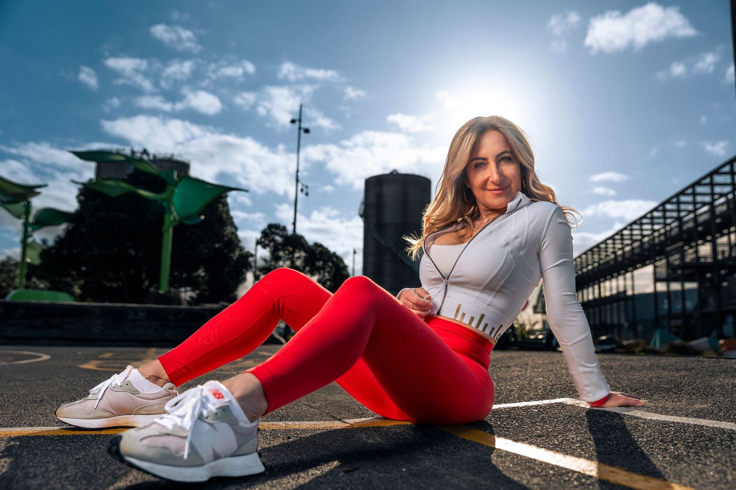 Woman in red leggings and grey crop top sitting on ground under a sunny blue sky.