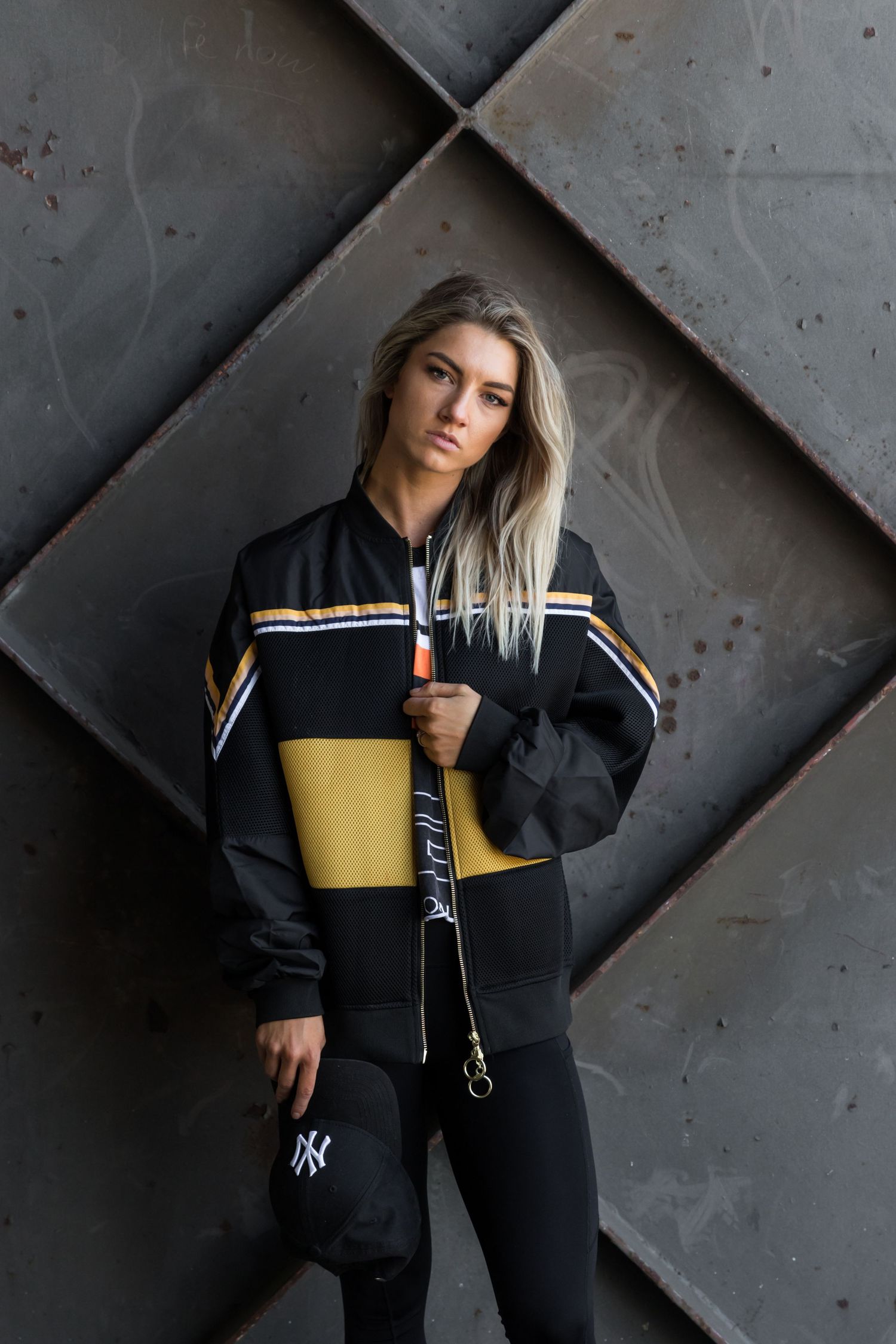 Blonde woman in black and gold NY Yankees tracksuit posing against a dark geometric metal wall.