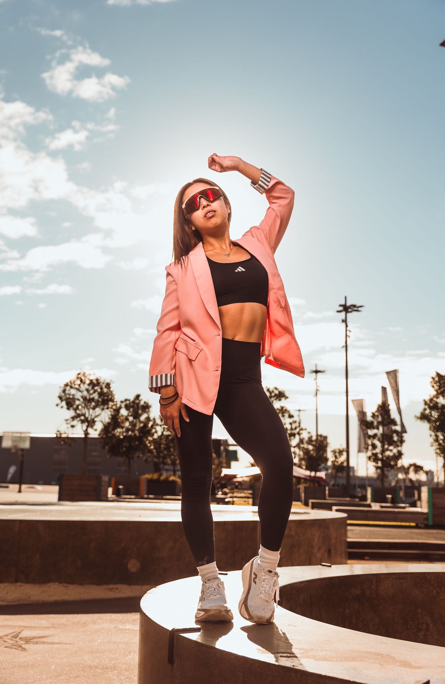 Woman in pink blazer and black Adidas activewear posing confidently outdoors on a sunny day.