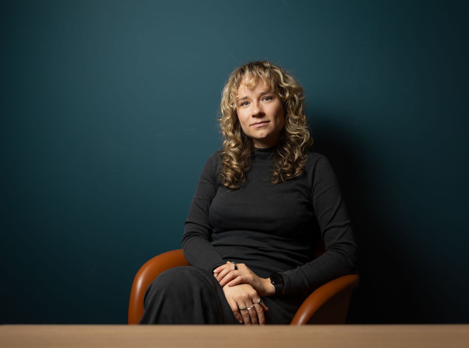 Woman with curly blonde hair sitting in a brown chair against a teal wall wearing a black turtleneck.