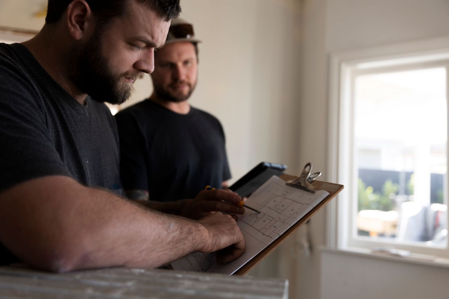 Two bearded construction workers studying blueprints on a clipboard inside a renovation site.