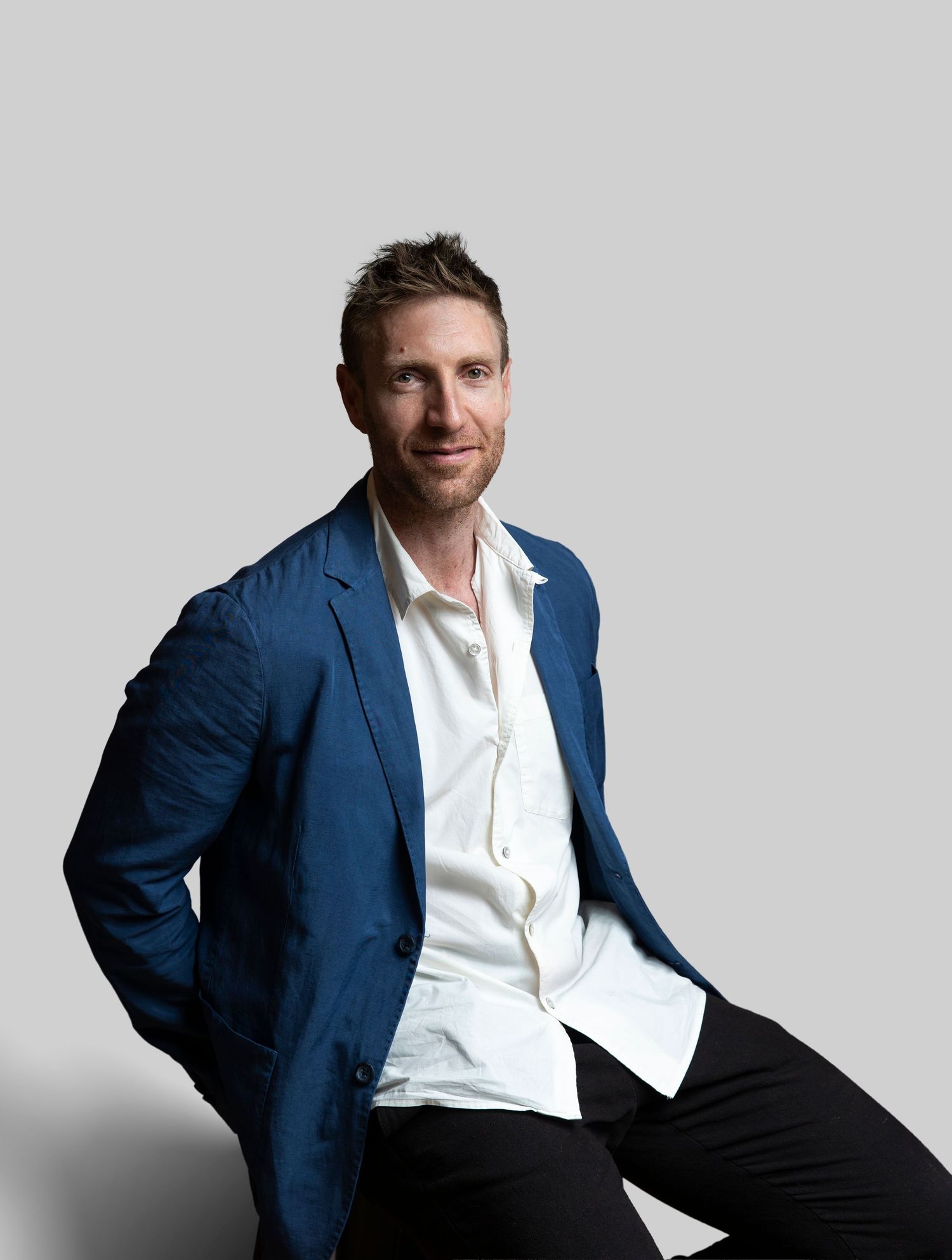 Man in blue blazer and white shirt sitting casually against a neutral gray background.