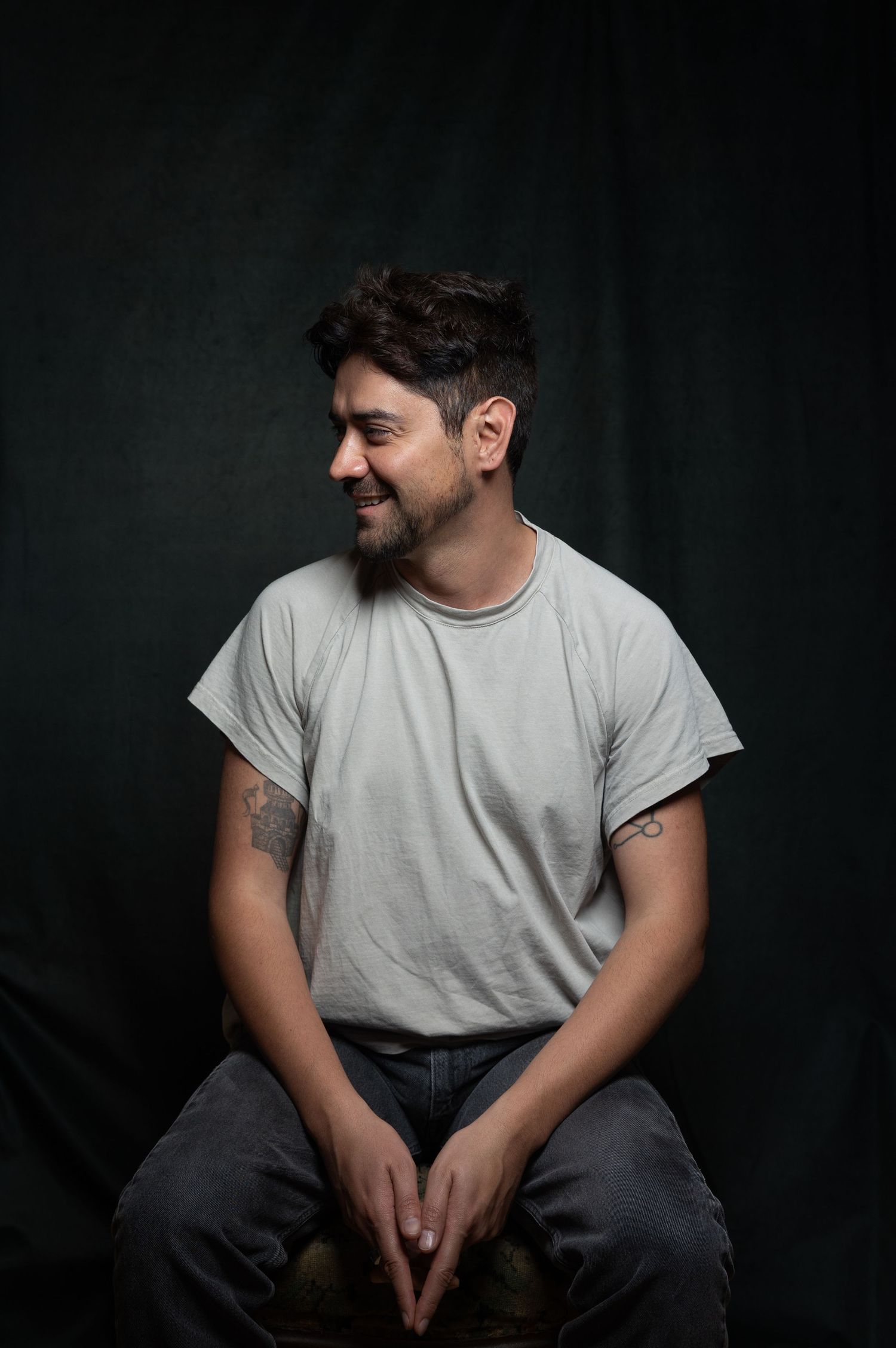 Bearded man in gray t-shirt sitting relaxed and smiling against a dark backdrop.