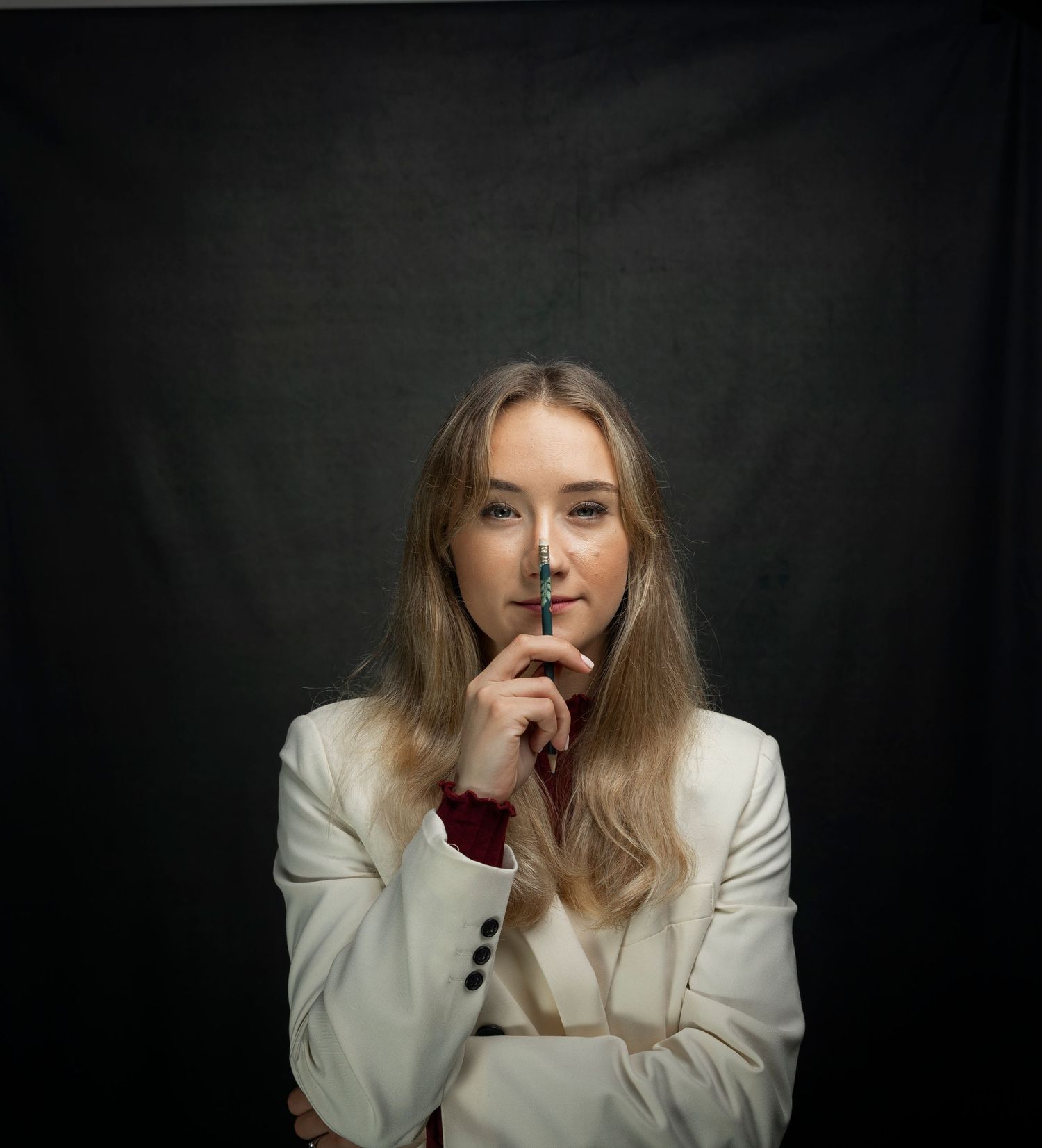 Young blonde woman in white blazer posing thoughtfully with finger to lips against dark background.