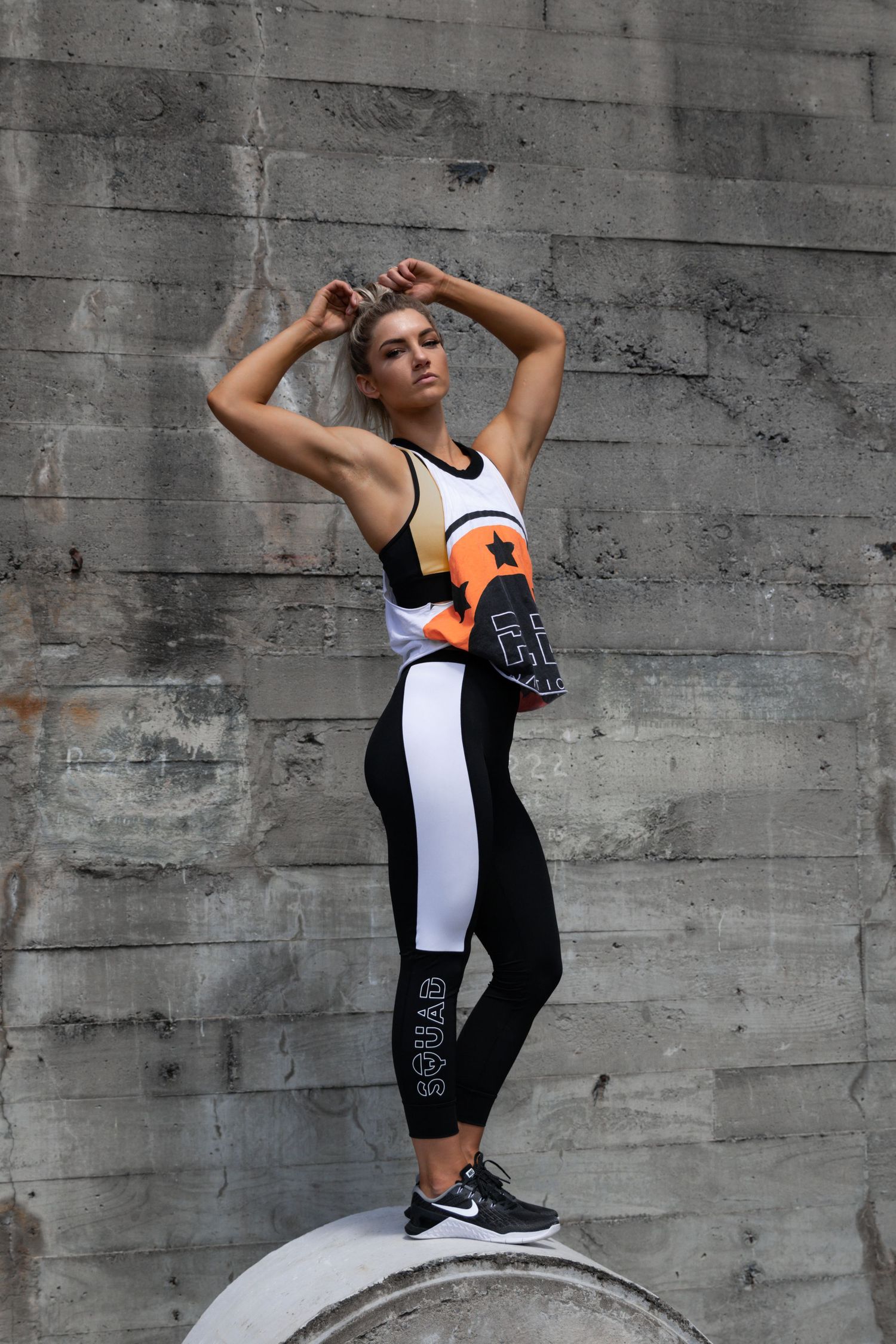 Athletic woman in orange and black fitness outfit standing on a concrete cylinder outdoors.