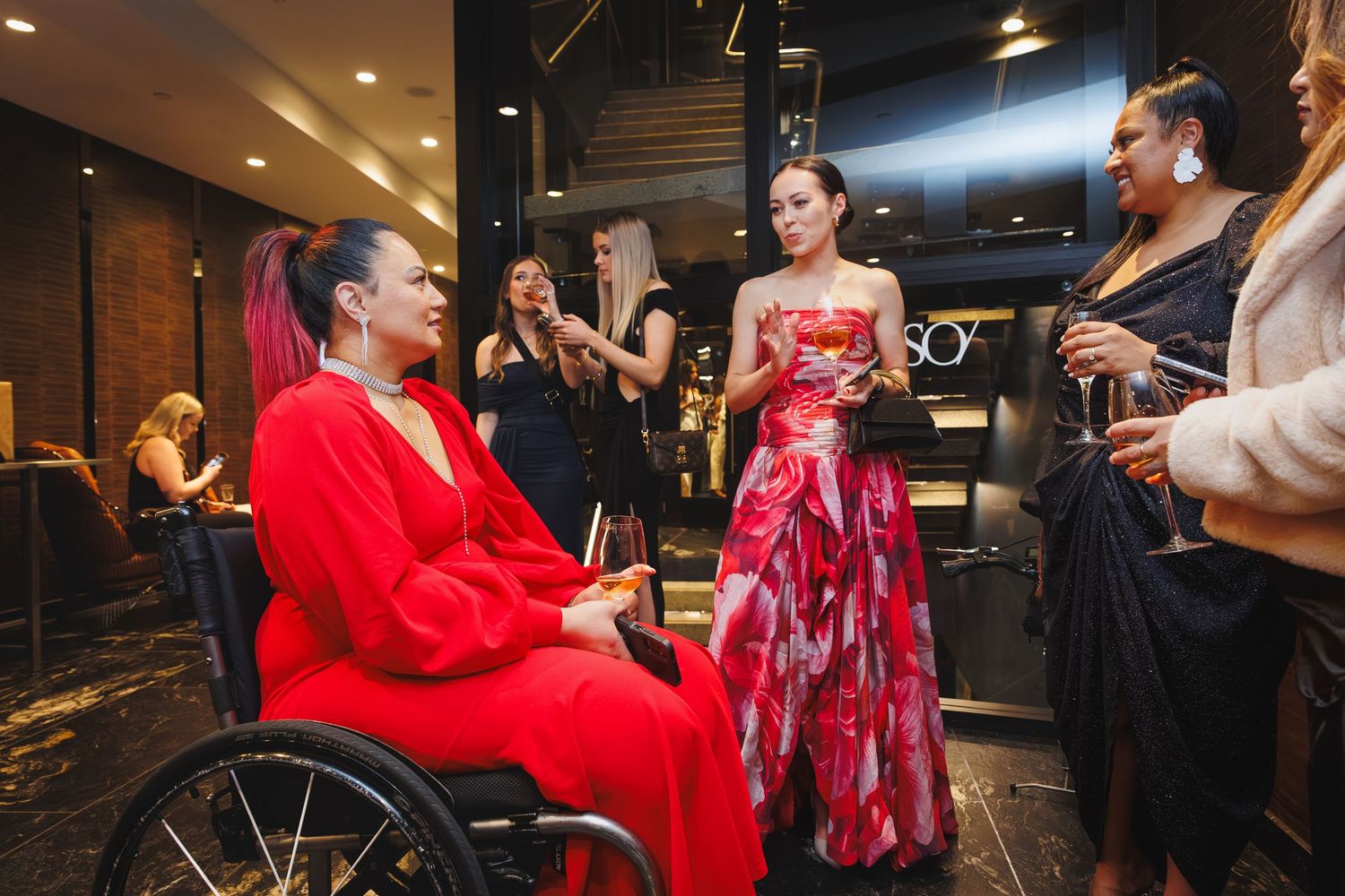 Women in formal attire socialize at an upscale event, including a woman in a red outfit in a wheelchair.