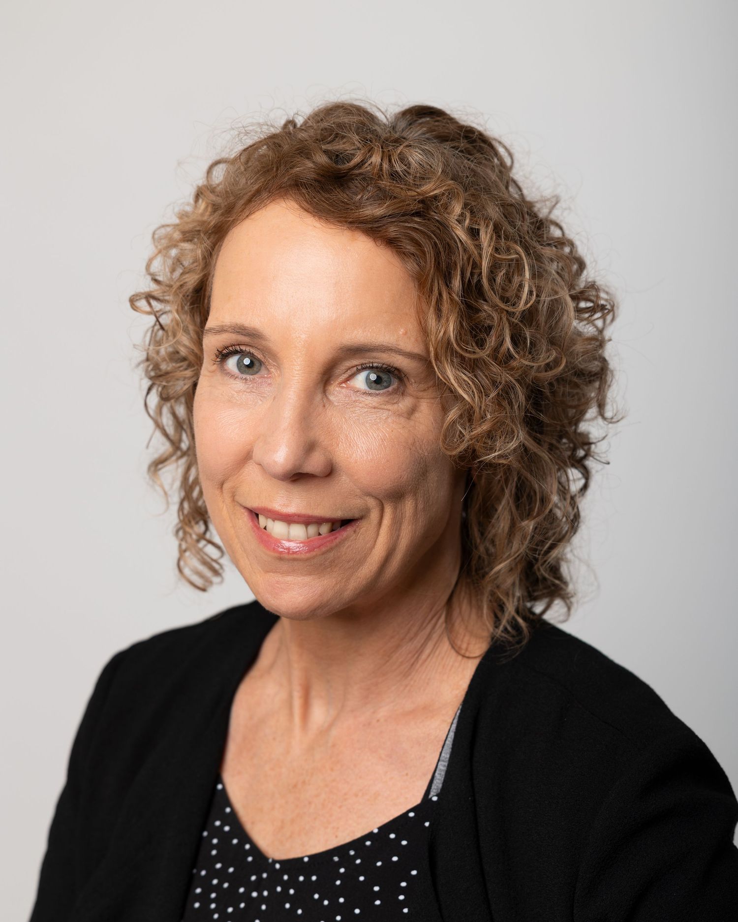 Professional headshot of a middle-aged woman with curly brown hair wearing a black cardigan and polka dot top.