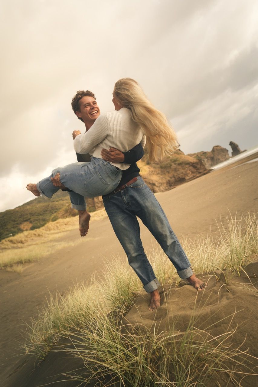 A joyful couple embracing on sandy beach dunes, man lifting woman as they laugh together under cloudy skies.