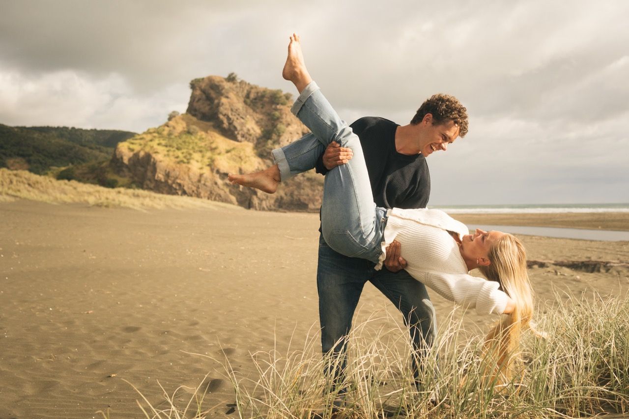 Couple dancing romantically on a sandy beach with dramatic rocky cliffs and cloudy skies in the background.