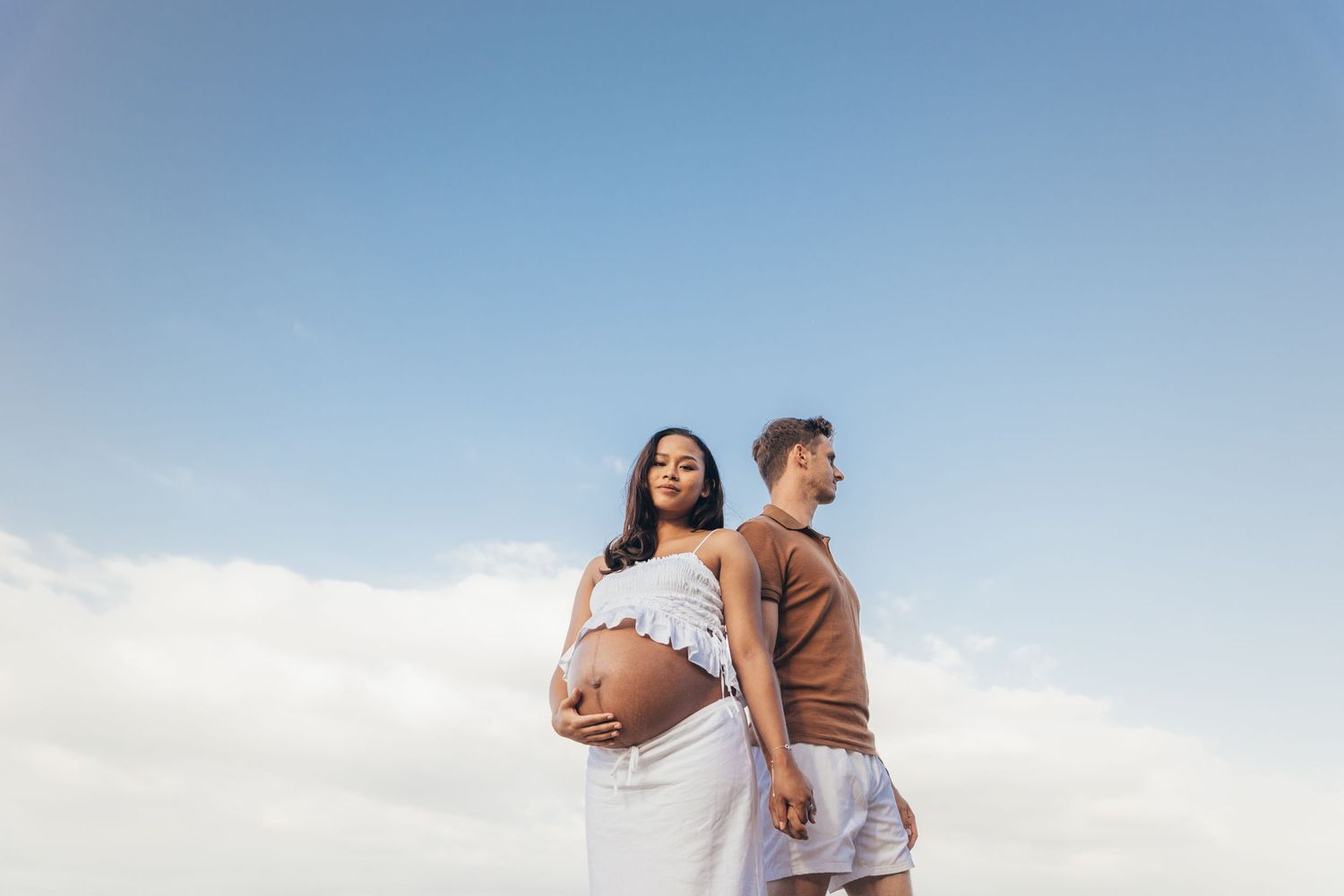 Pregnant woman in white dress stands back-to-back with partner under blue sky, cradling her baby bump.