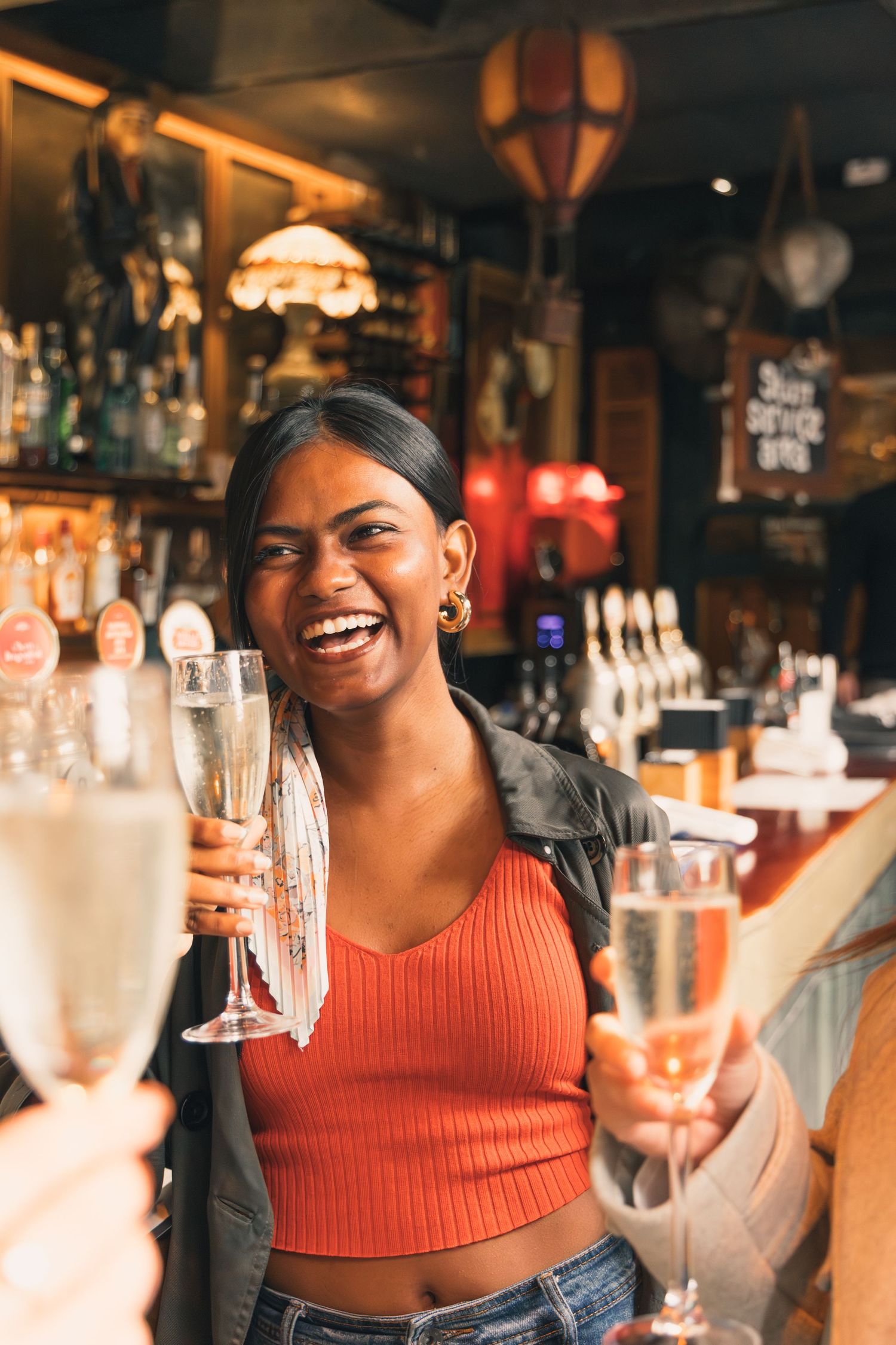 A smiling young woman toasting with champagne glasses at a bar, wearing an orange crop top and jacket.
