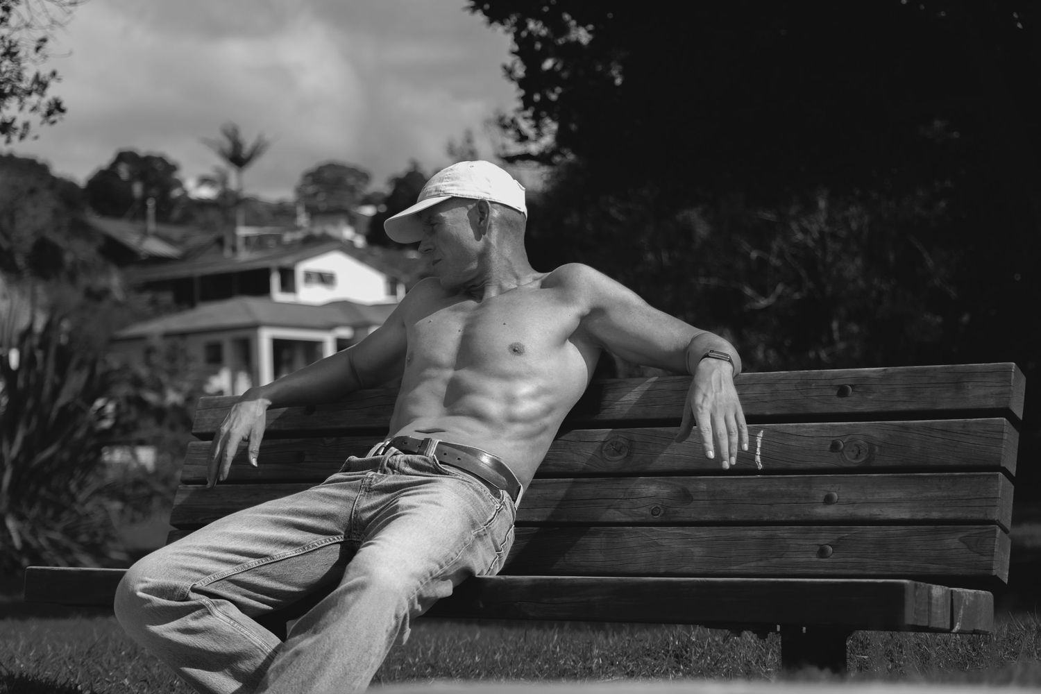 Fit shirtless man relaxing on a park bench wearing a white cap and jeans in a black and white photo.