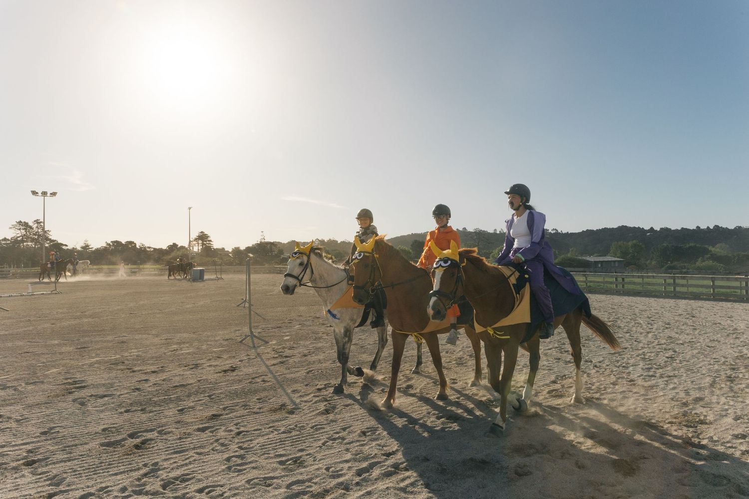 Two young riders on brown horses walking through a sunny sandy equestrian training arena.
