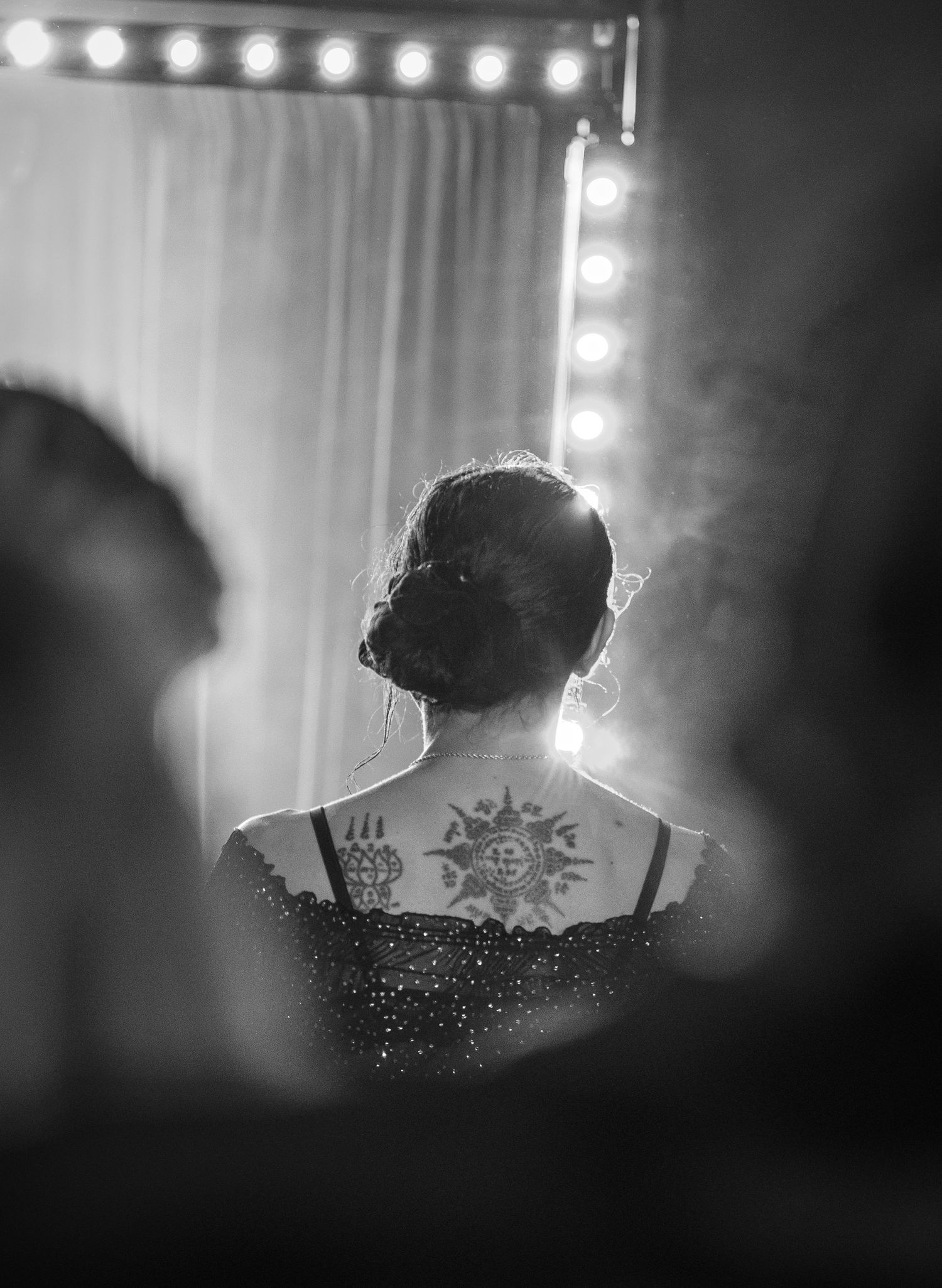 Black and white back view of woman with sun tattoo and updo hair near vanity lights.