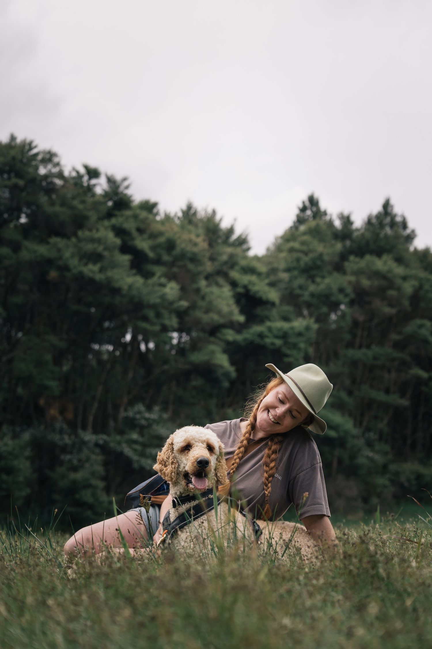 Smiling woman in hat sitting in grass field with golden labradoodle dog on leash with forest background.