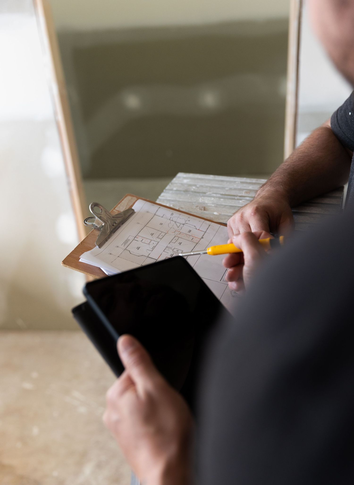 Builder holding tablet and pencil reviewing floor plan blueprints on a clipboard.