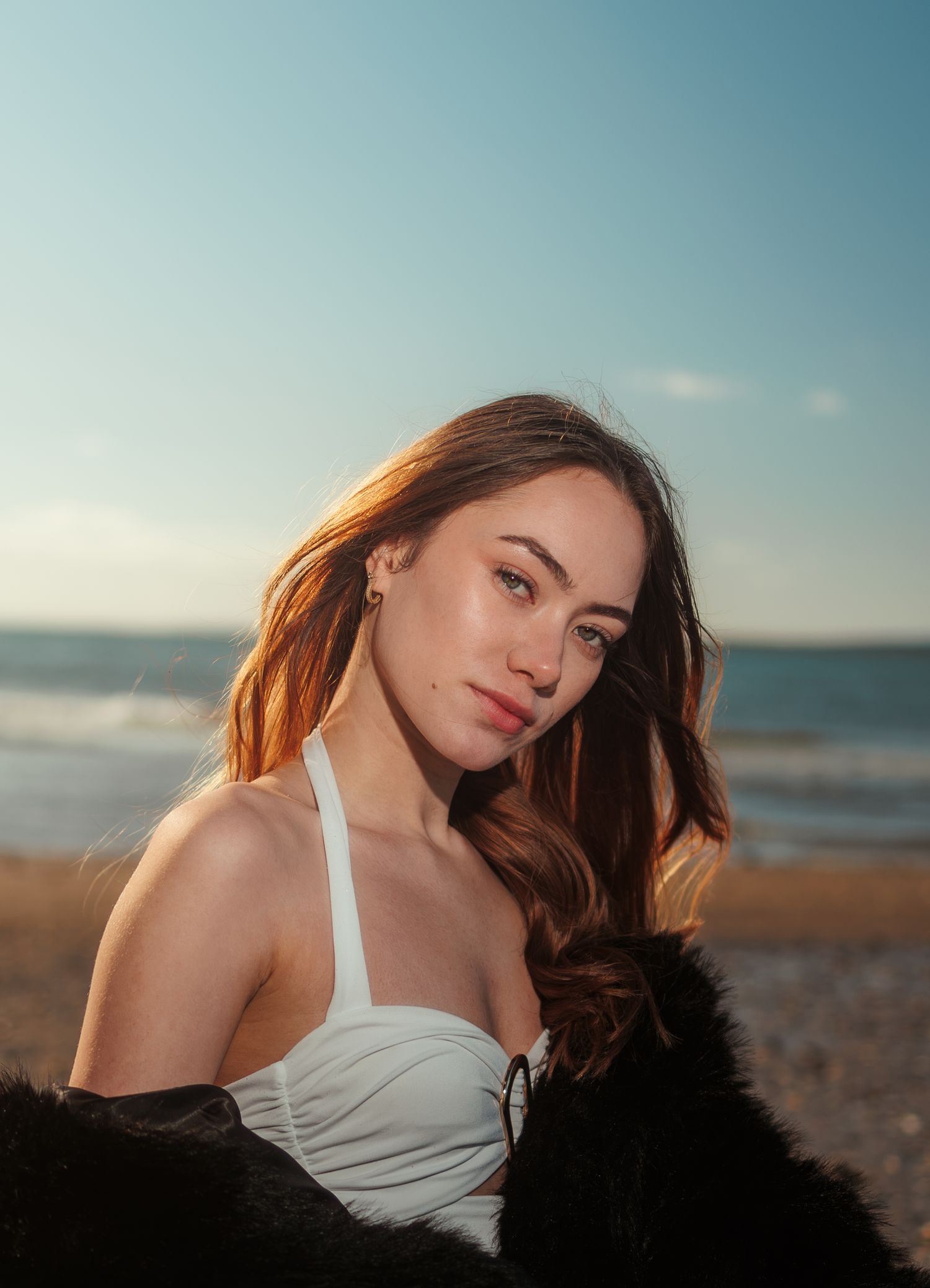 Young woman with auburn hair in white swimsuit and black fur coat poses on a sunny beach at golden hour.