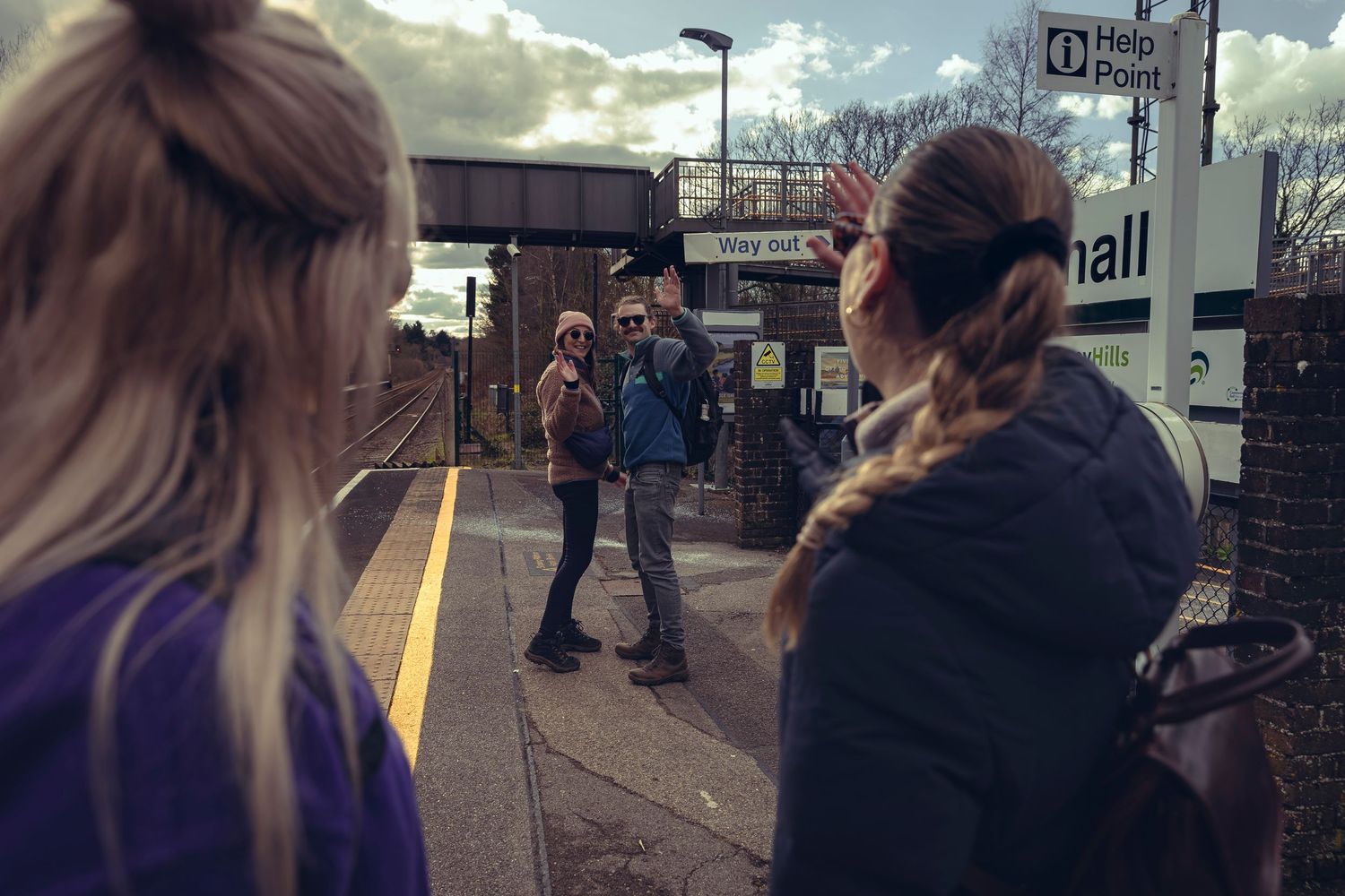 People waiting on a suburban train station platform on a cloudy day.