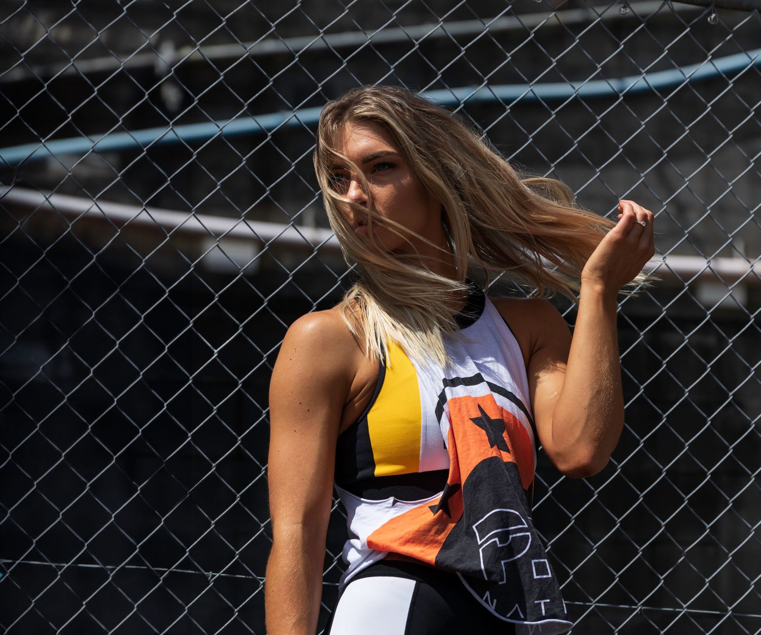 Blonde athletic woman in colorful sports outfit posing near a chain-link fence outdoors.