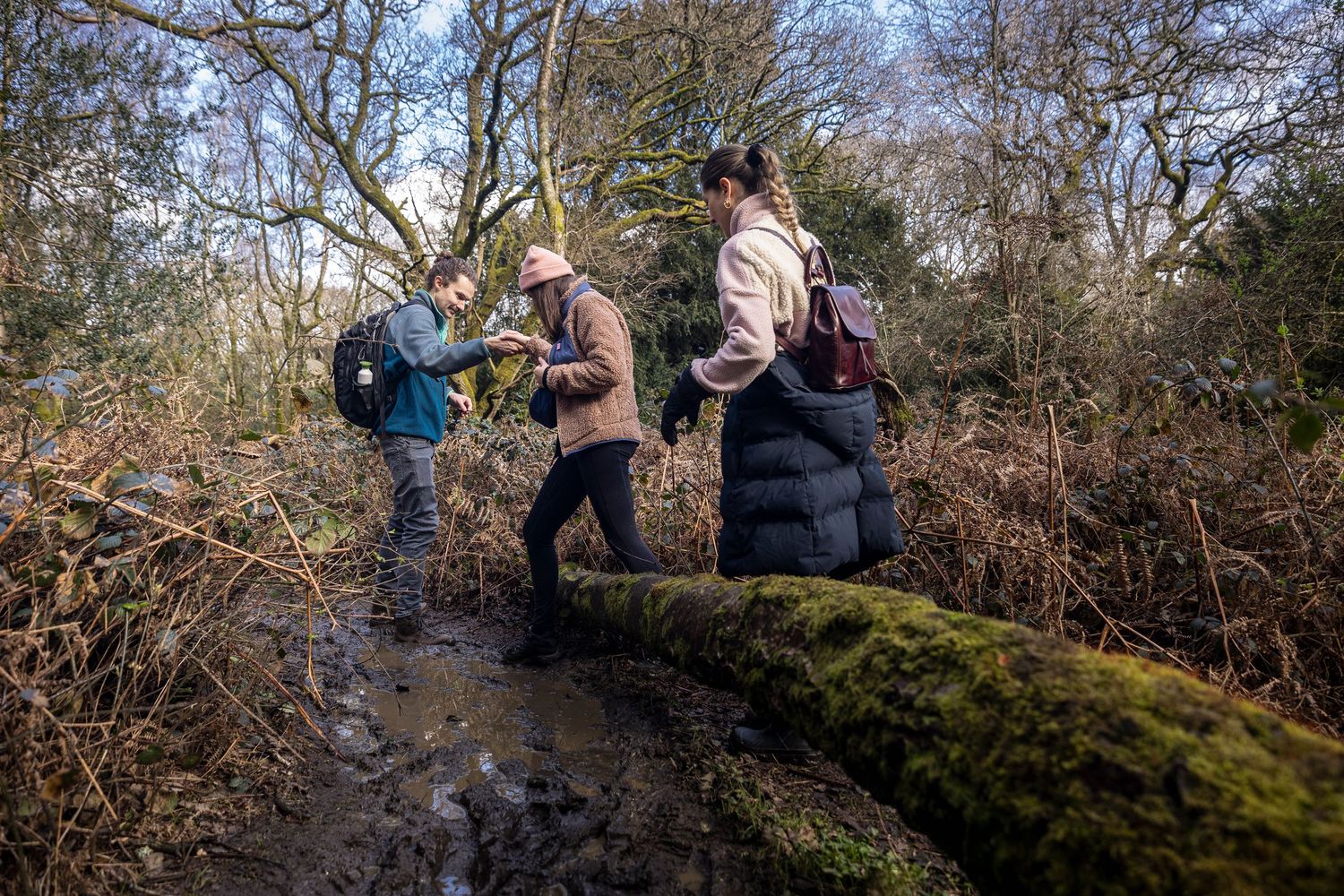 Three hikers crossing a mossy log on a muddy woodland nature trail.